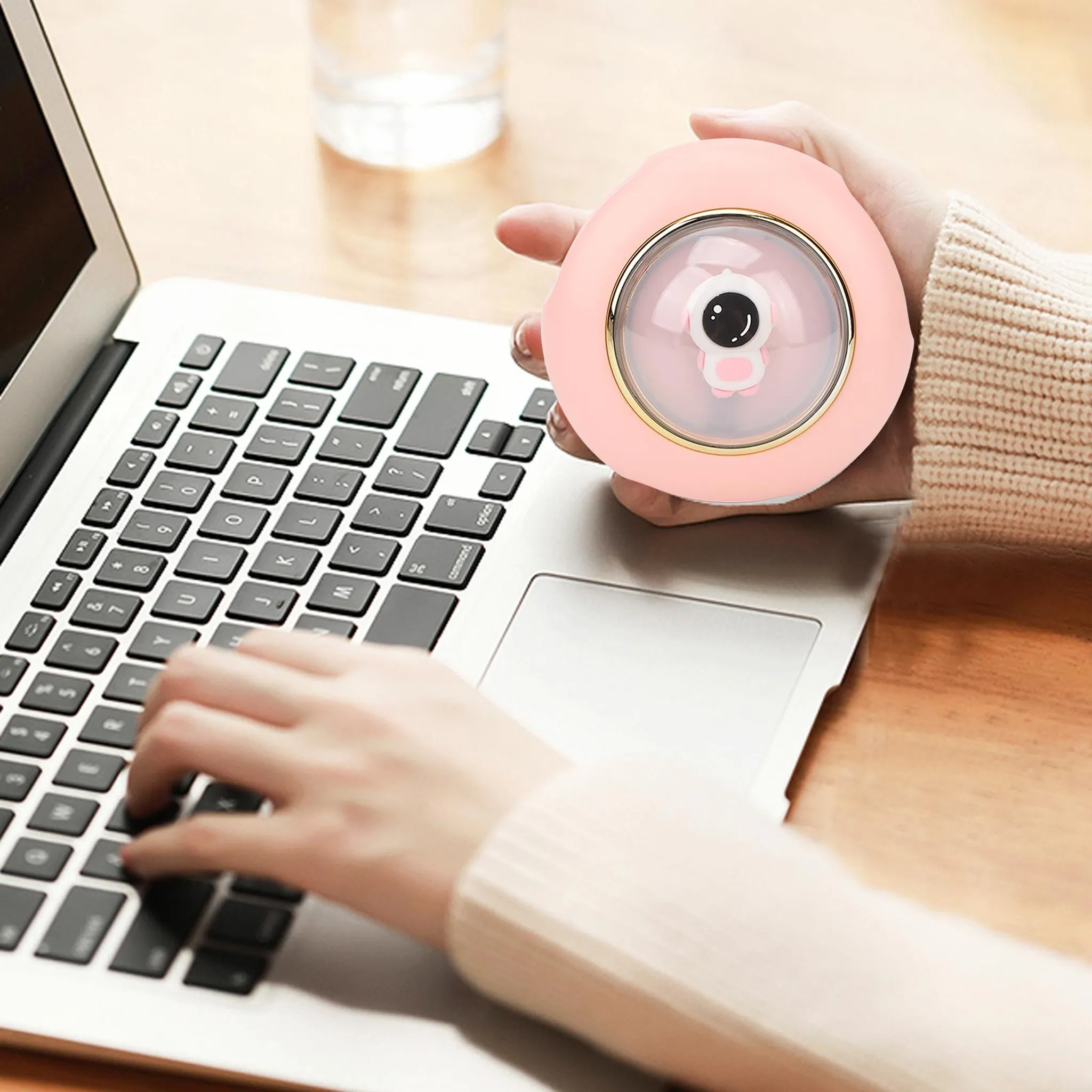 Eine Person benutzt einen Laptop. Sie halten einen rosa Handwärmer in Form einer Cartoonfigur. Im Hintergrund steht ein Glas.