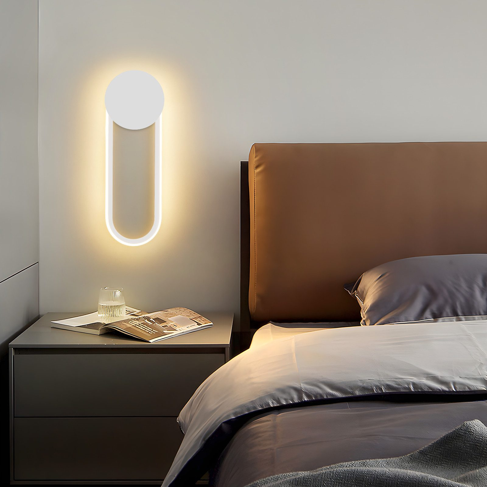Scena di camera da letto con lampada da parete illuminata, comodino con bicchiere e rivista e un letto.