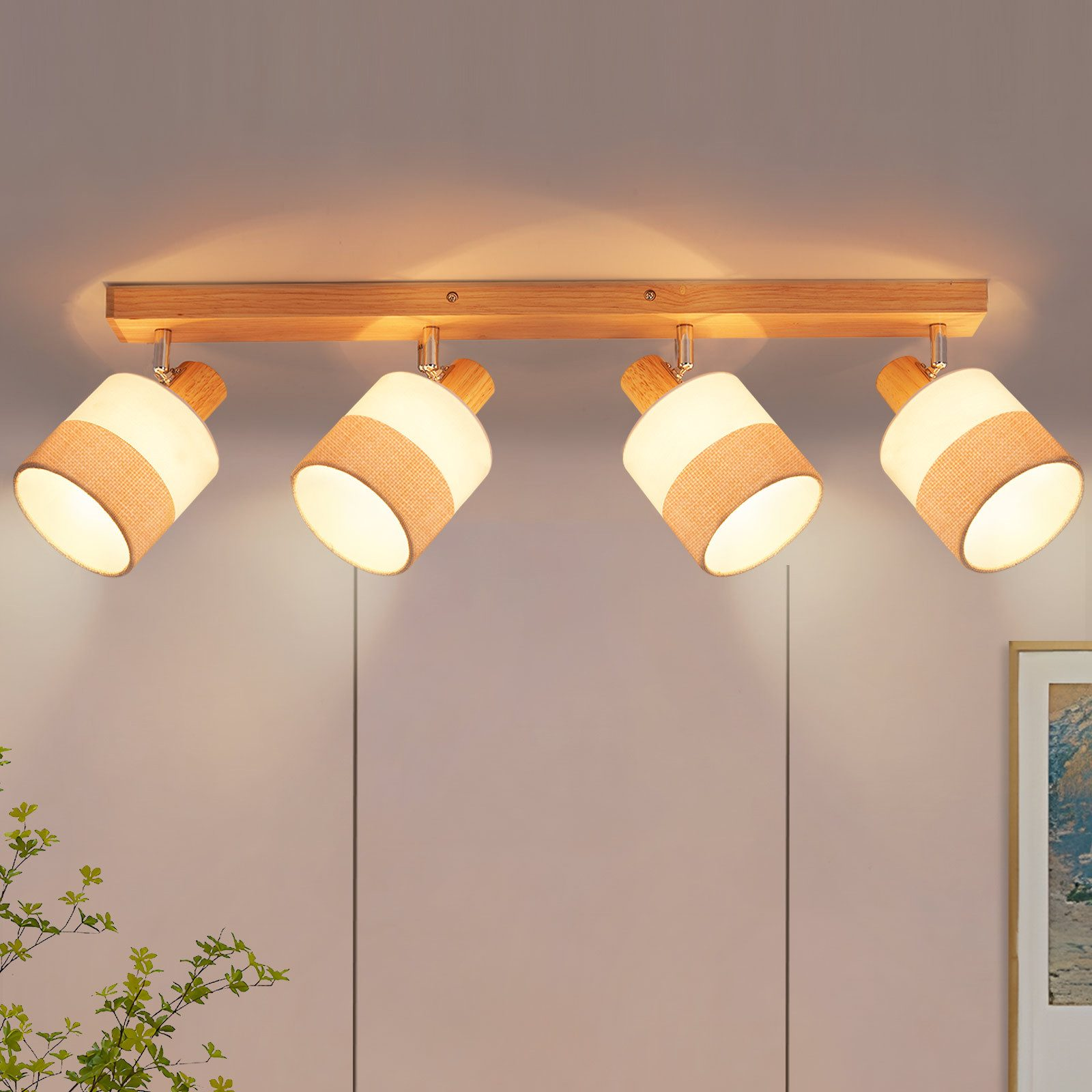 Quattro faretti su una trave di legno. Ognuno ha un paralume bianco con una fascia beige. Lo sfondo è un muro con una pianta.
