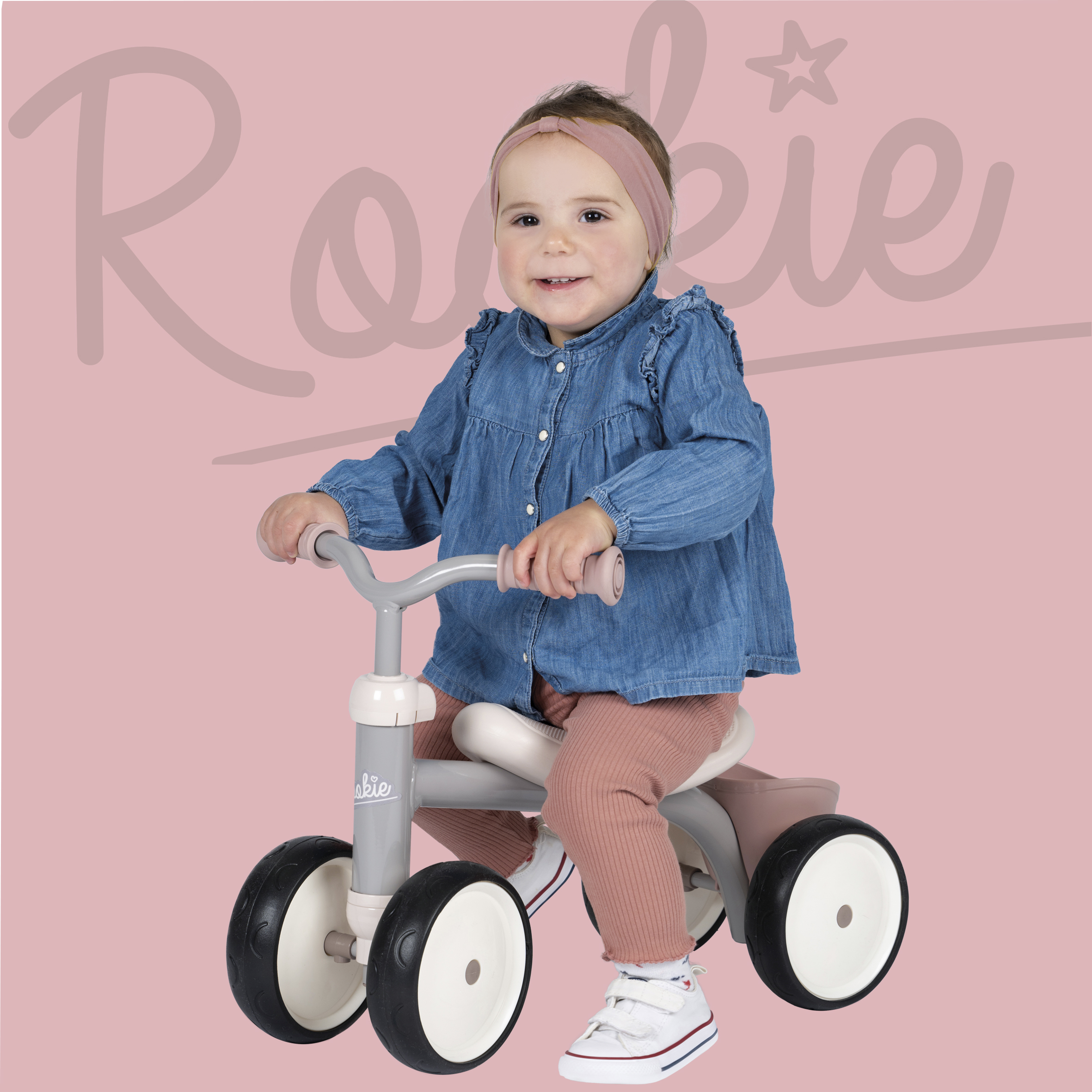Ein Baby fährt Dreirad. Das Baby trägt ein Jeanshemd, eine rosa Hose und weiße Schuhe. Rosa Hintergrund.