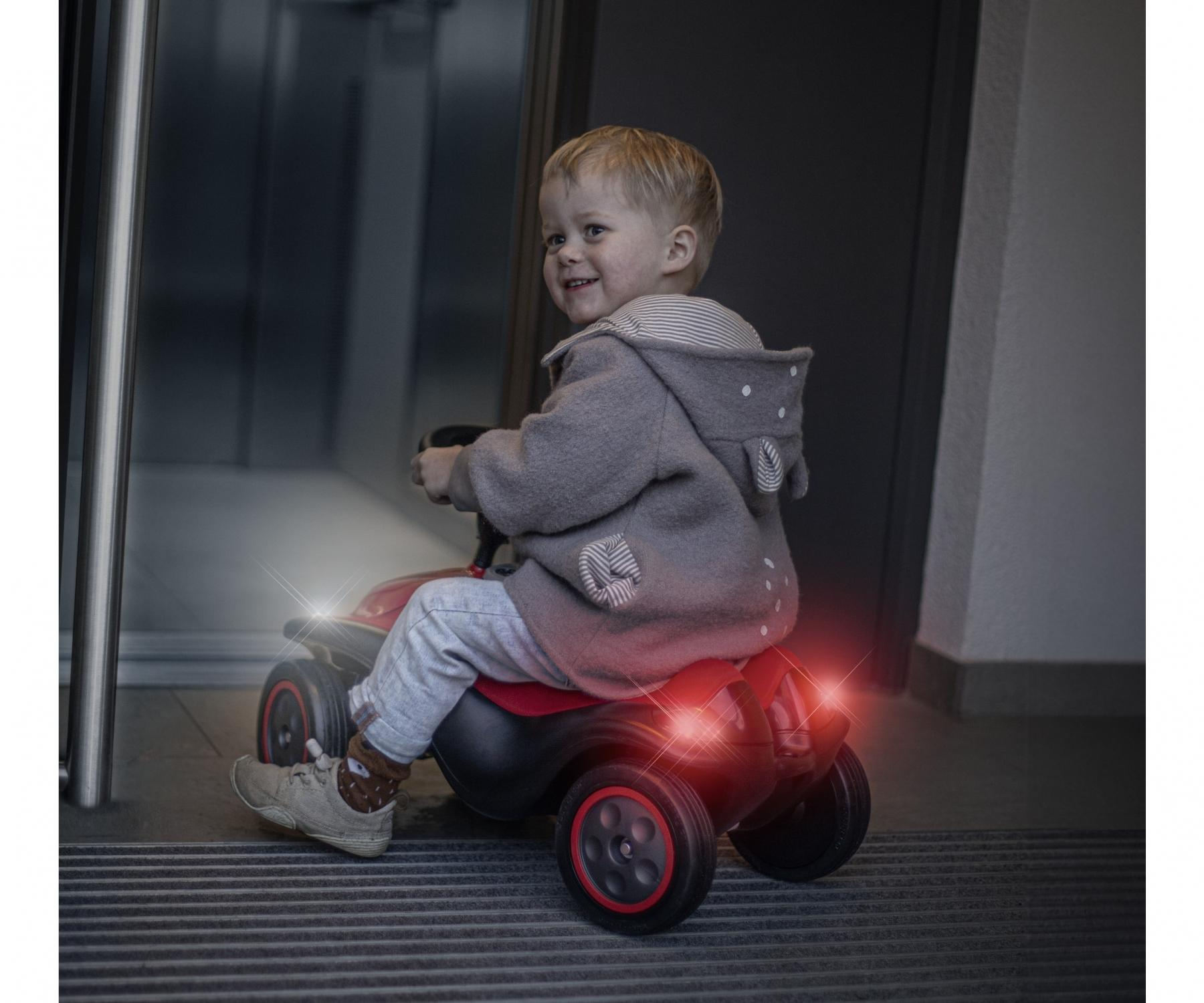Ein Kind in einem grauen Mantel fährt mit einem roten Spielzeugauto. Das Auto hat Scheinwerfer und Rücklichter. Das Kind lächelt.