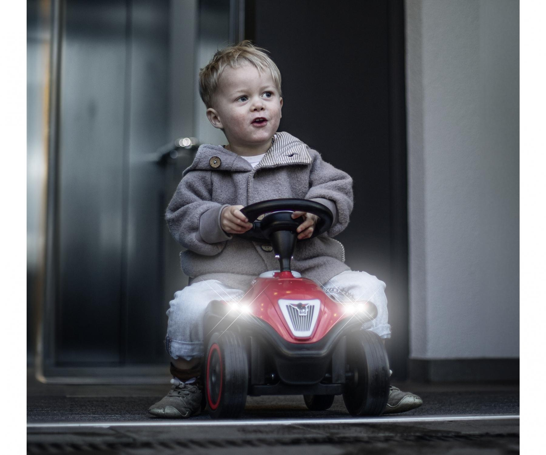 Ein kleiner Junge in einem grauen Mantel sitzt auf einem roten Spielzeugauto. Er hält das Lenkrad. Die Scheinwerfer des Autos sind an.
