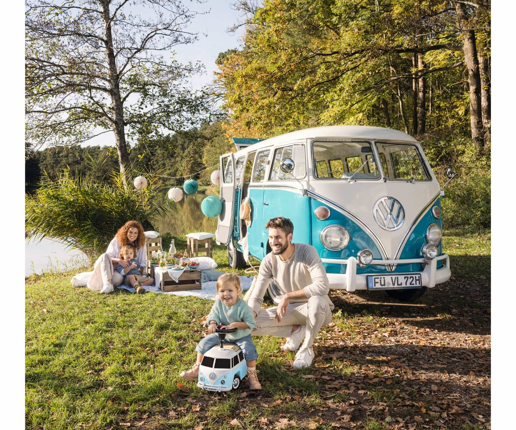 Familienpicknick in der Nähe eines Sees mit einem klassischen blau-weißen VW-Bus. Ein Kind fährt einen Spielzeug-Van, andere genießen ein Picknick.
