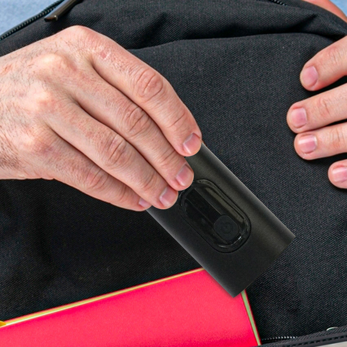 Hände halten ein schwarzes Gerät neben einer schwarzen Tasche. Rotes Notizbuch ragt aus der Tasche.
