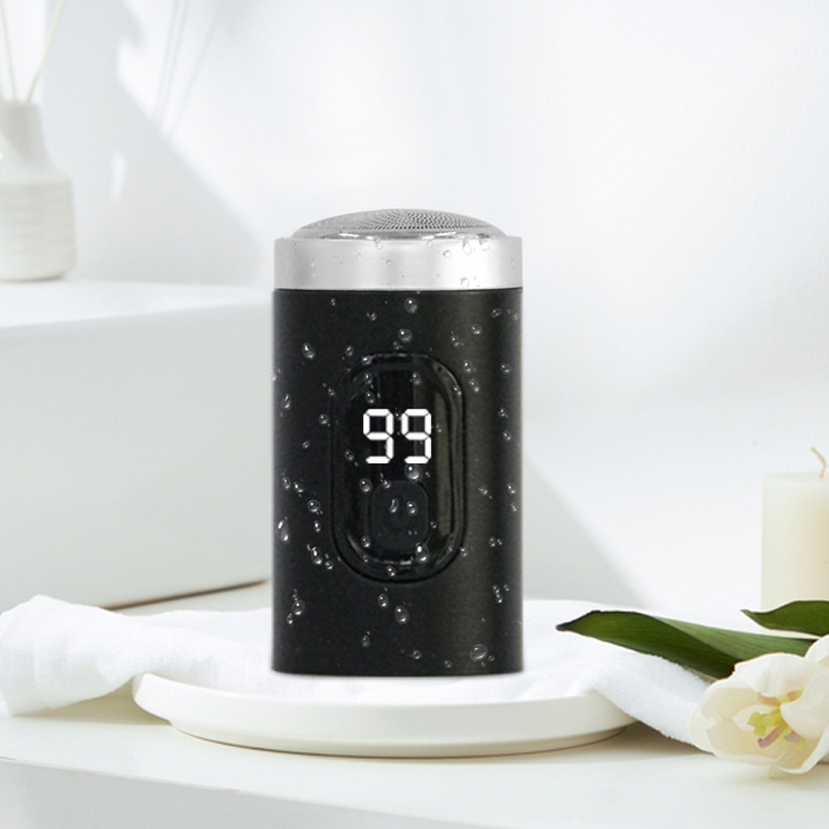 Ein schwarzer Elektrorasierer mit digitaler Anzeige '99' steht auf einem weißen Teller mit Wassertropfen. Rechts befindet sich eine weiße Blume.