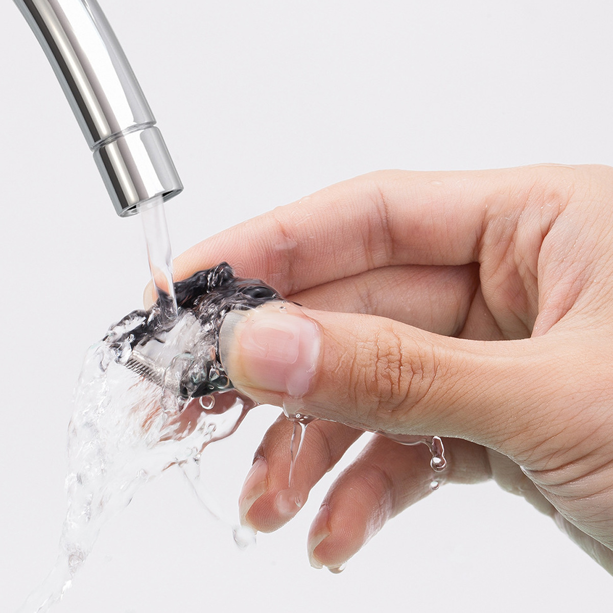 Eine Hand hält einen Rasiererkopf unter fließendem Wasser aus einem Metallhahn. Wassertropfen sind vor weißem Hintergrund sichtbar.