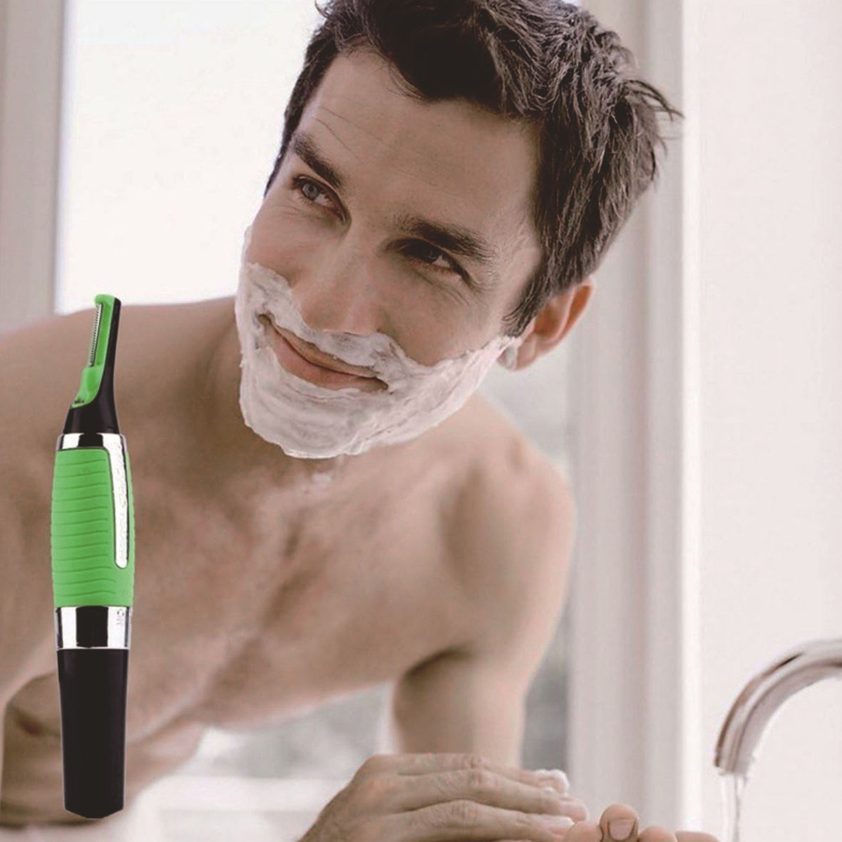 Mann mit Rasiercreme im Gesicht, blickt in den Spiegel. Ein grüner und schwarzer Trimmer ist im Vordergrund. Im Hintergrund ist ein Wasserhahn.