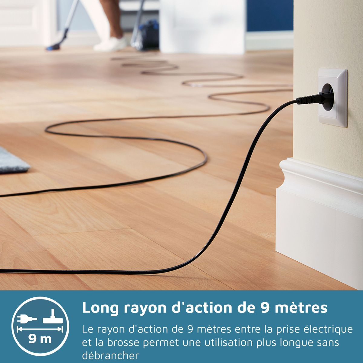 Kabel od odkurzacza wpięty do gniazdka ściennego, rozciągający się po drewnianej podłodze. Osoba odkurza w tle.