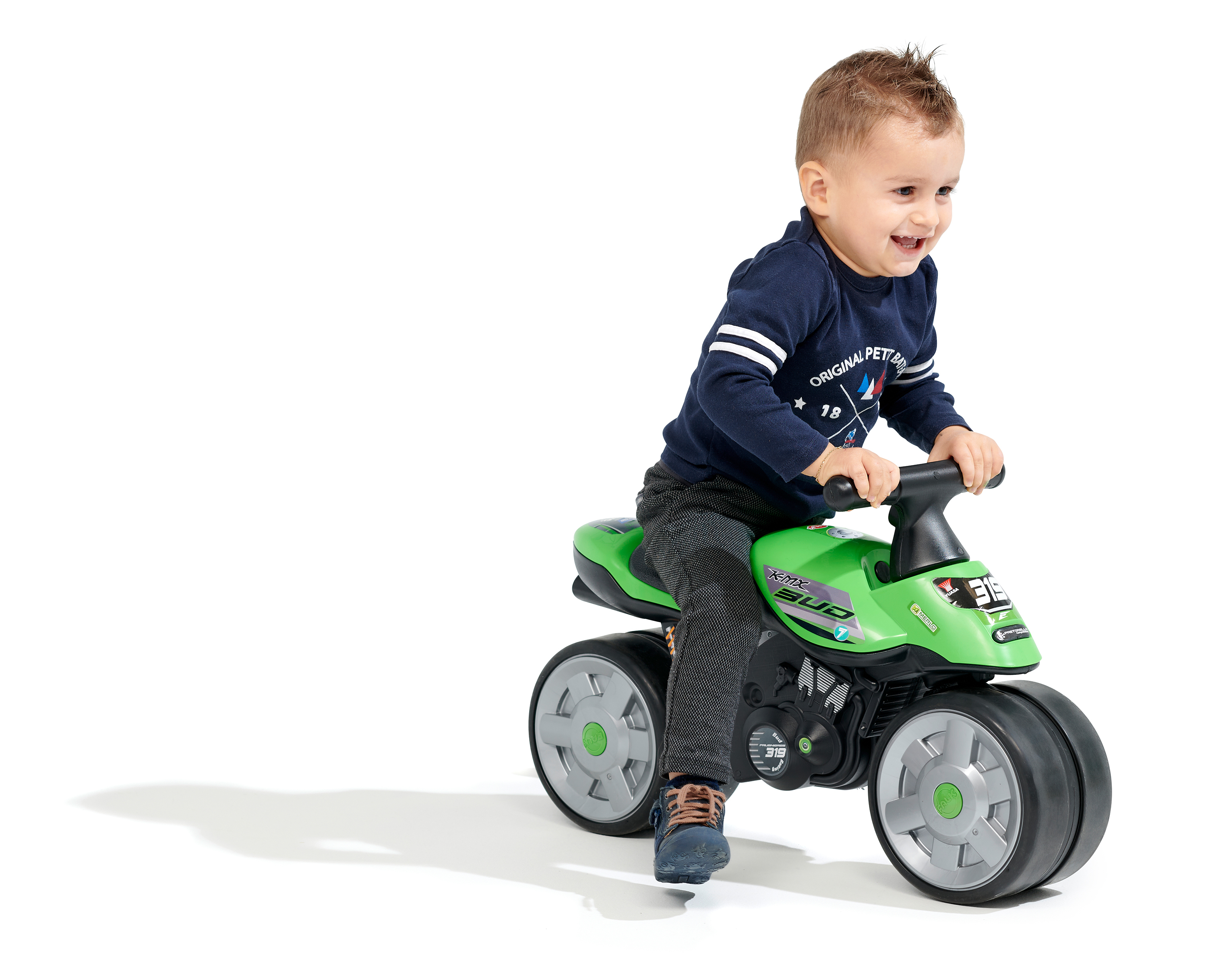 Un bambino sorridente cavalca una moto giocattolo verde su uno sfondo bianco. Indossa una camicia blu e pantaloni grigi.