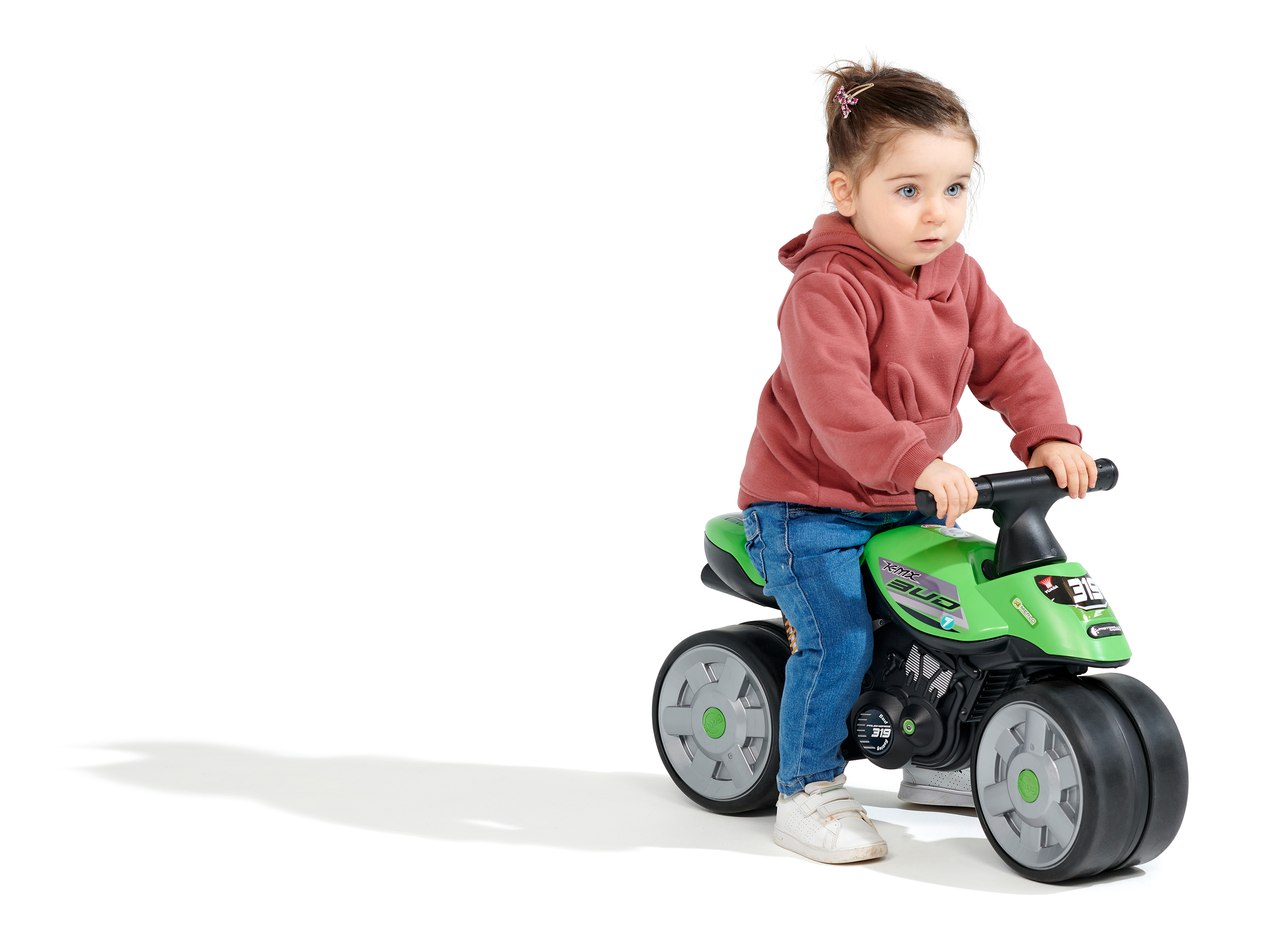 Un bambino piccolo su una moto giocattolo verde, indossa una felpa con cappuccio rossa e jeans, su sfondo bianco.