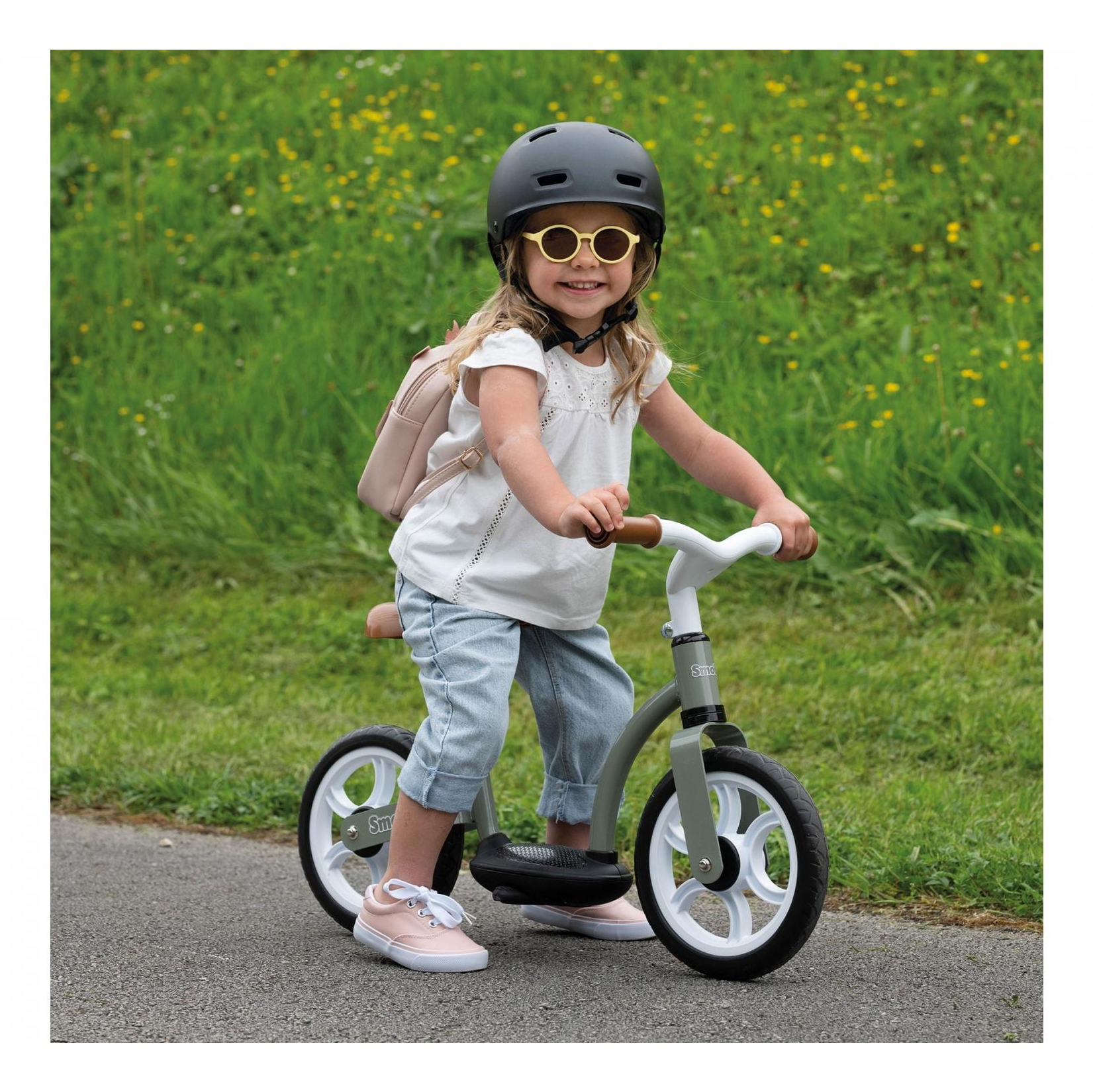 Una bambina su una bici senza pedali con casco e occhiali da sole, sorridente. Lo sfondo è un campo erboso con fiori gialli.