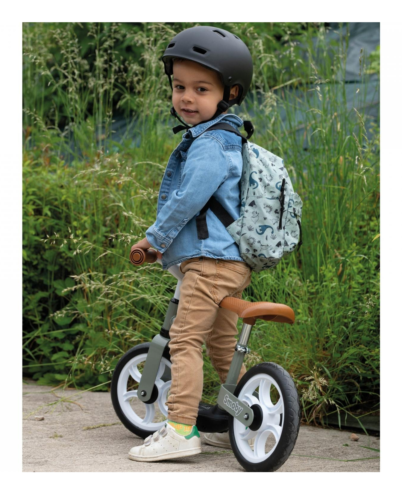 Un bambino con casco e zaino va in bici senza pedali. Erba verde sullo sfondo. Il bambino indossa una giacca di jeans e pantaloni marroni.