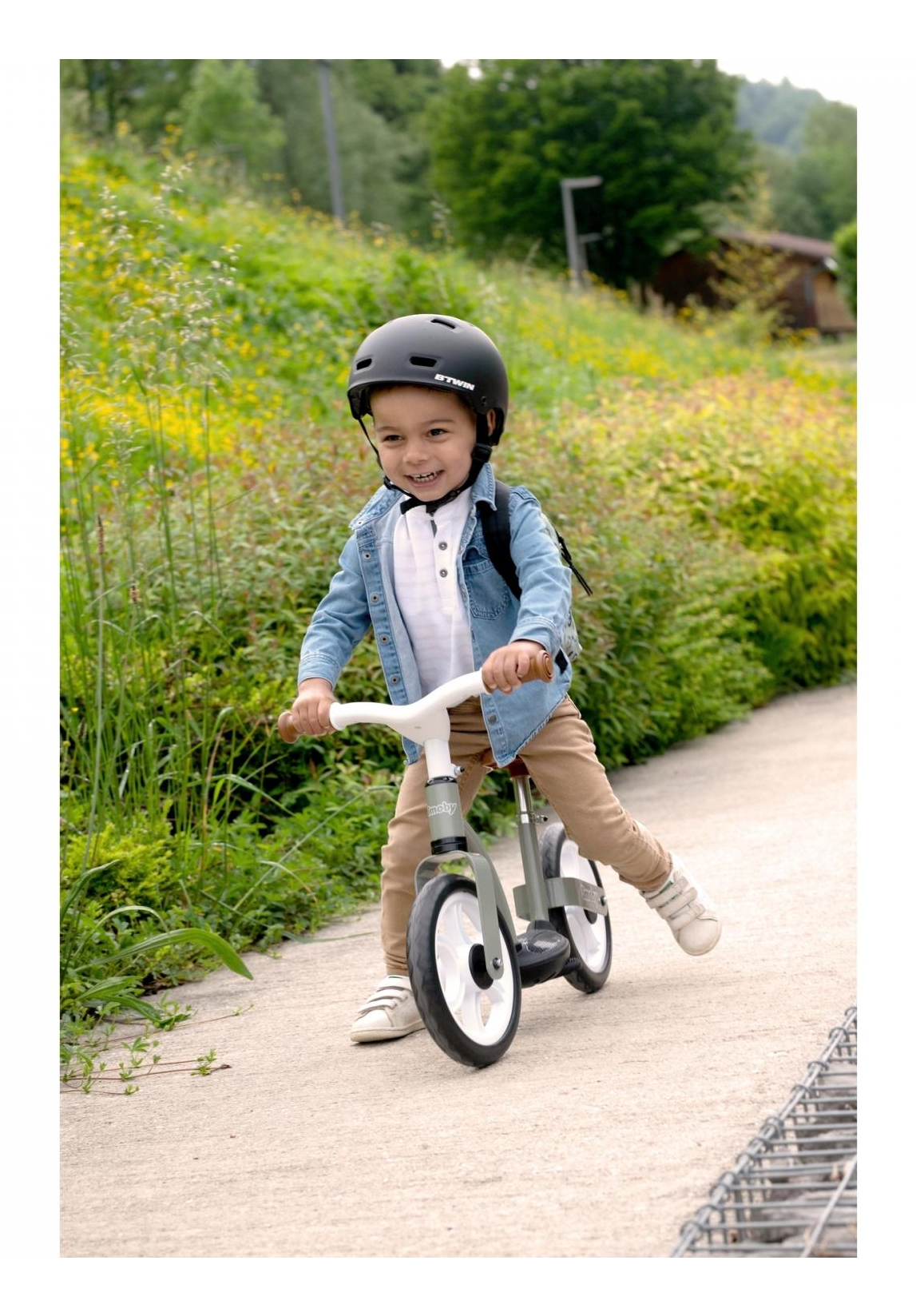 Un bambino con un casco nero va in bici senza pedali su un sentiero. Il bambino sorride, circondato da verde e alberi sullo sfondo.