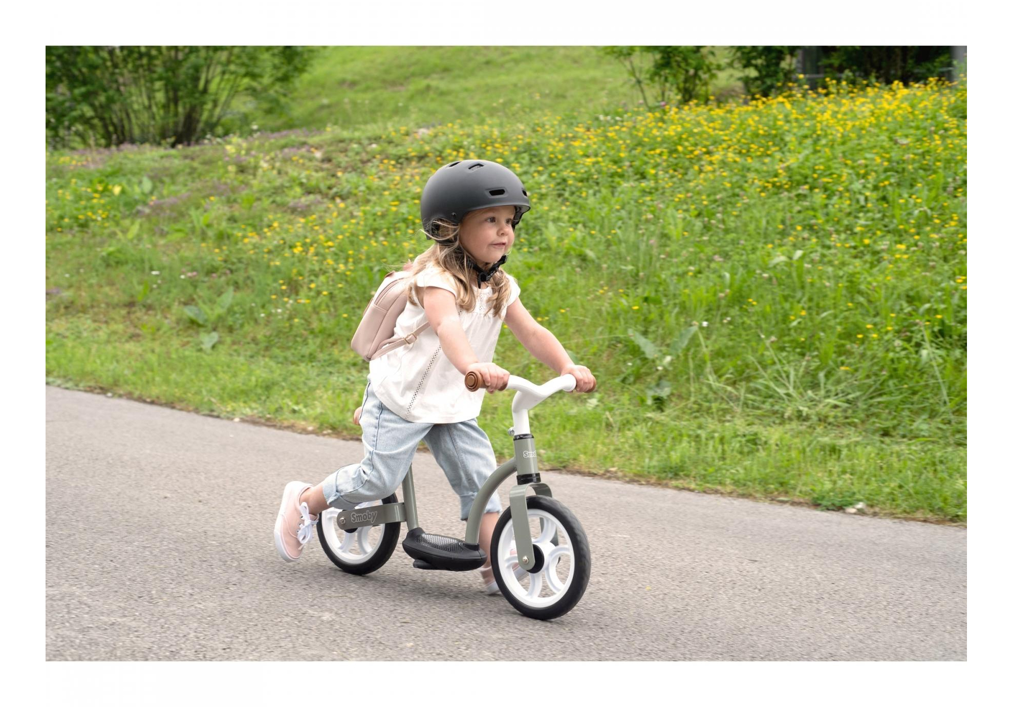 Una bambina con un casco cavalca una bici senza pedali su un sentiero, con erba e fiori sullo sfondo.