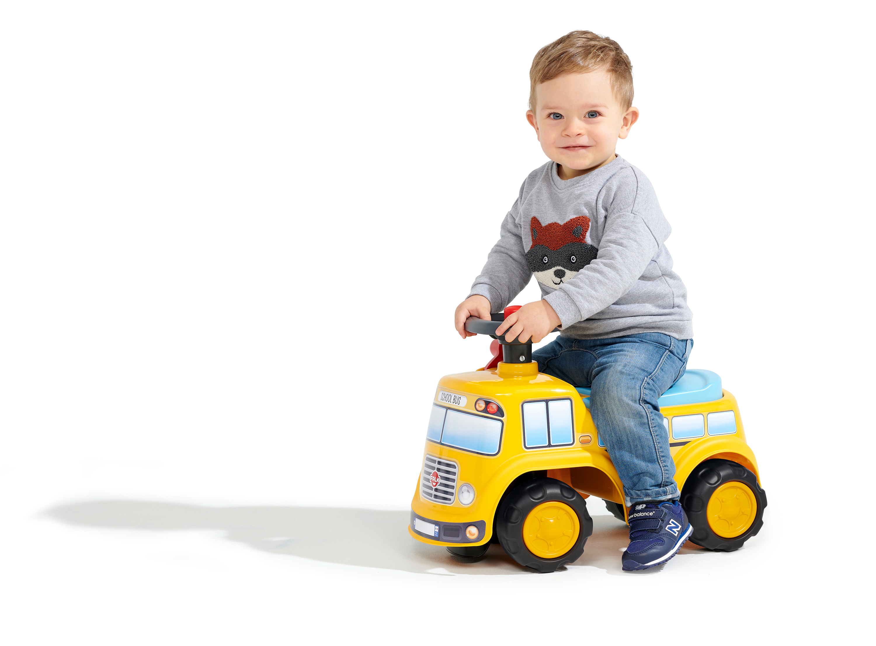 Ein lächelndes Kleinkind fährt mit einem gelben Schulbus-Spielzeug. Er trägt Jeans, einen grauen Pullover und blaue Schuhe, vor weißem Hintergrund.