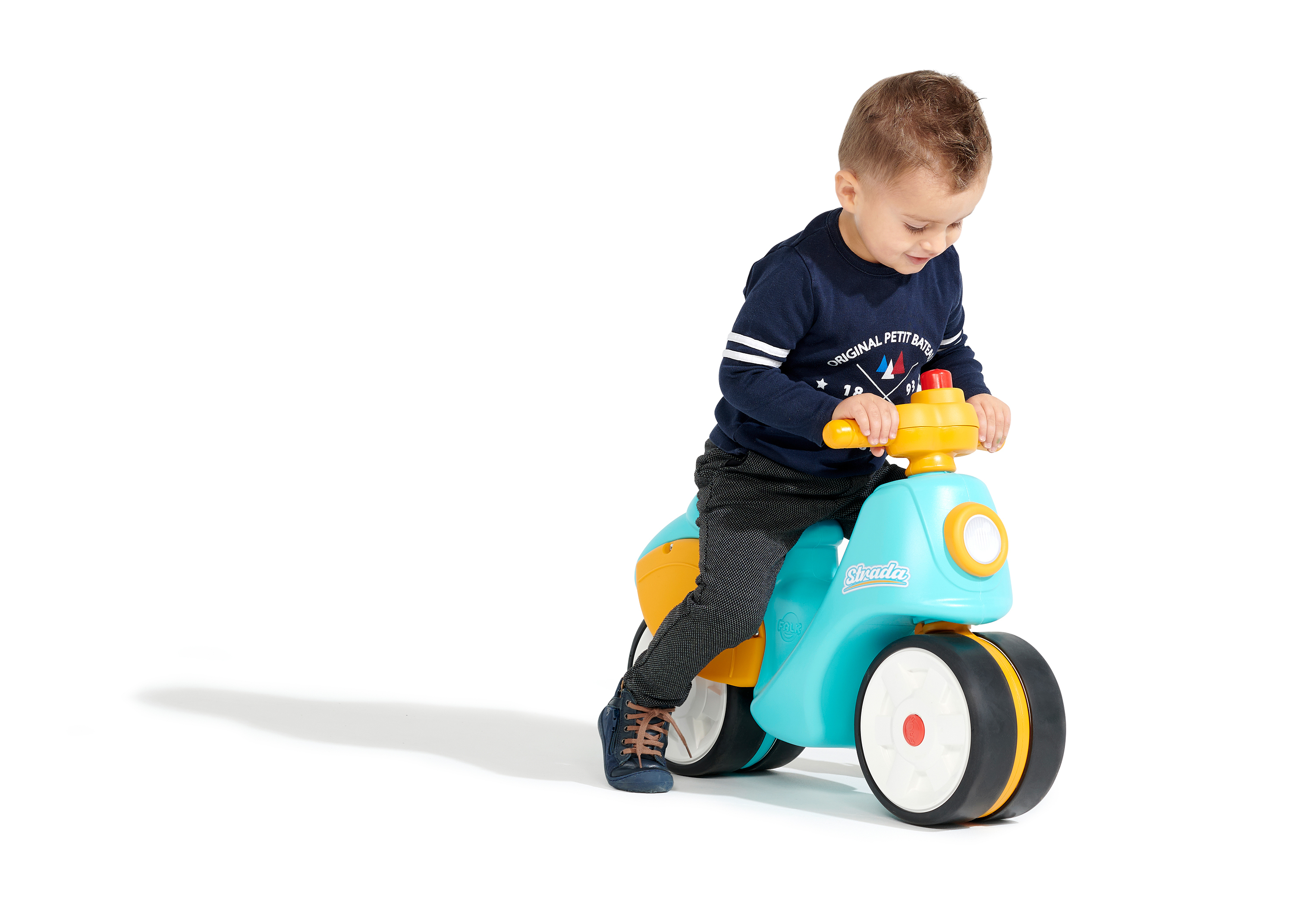 Ein kleiner Junge sitzt auf einem blau-gelben Spielzeugmotorrad. Er trägt ein marineblaues Hemd und graue Hosen. Der Hintergrund ist weiß.