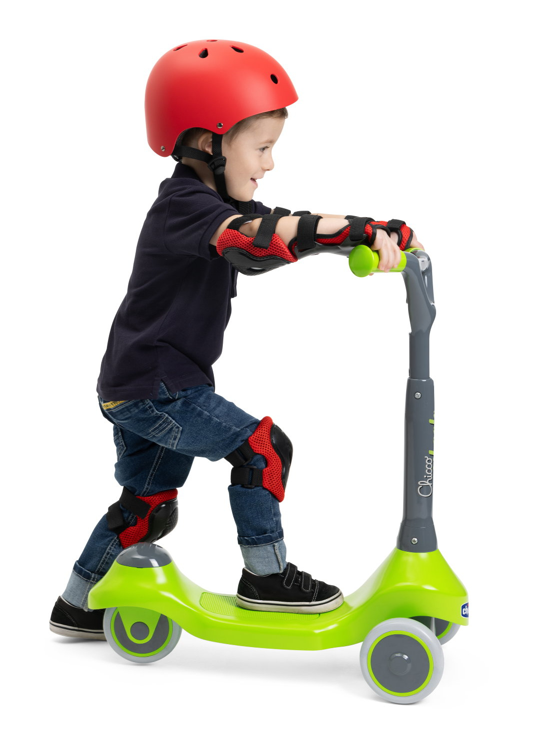 Un bambino con casco rosso e protezioni guida un monopattino verde. Il bambino indossa jeans e scarpe nere.