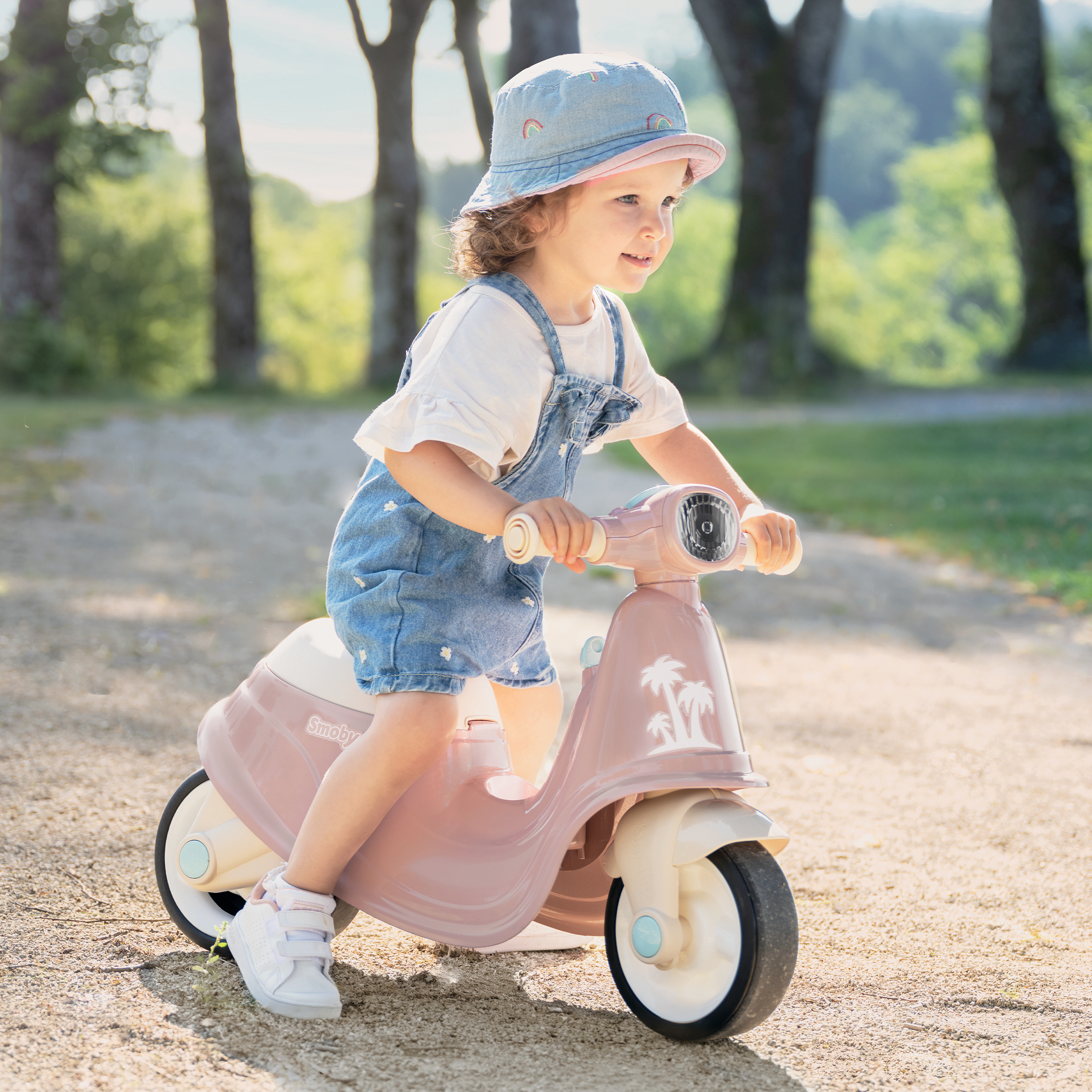 Ein Kleinkind fährt mit einem rosa Roller auf einem Kiesweg in einem Park. Sie trägt ein Jeans-Outfit und einen Hut mit Regenbogen.