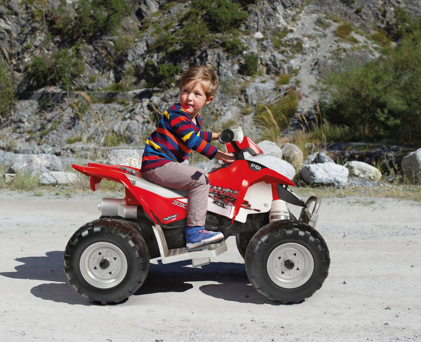Un ragazzino guida un quad rosso su una strada sterrata. Indossa una camicia a righe. Lo sfondo ha rocce e alberi.