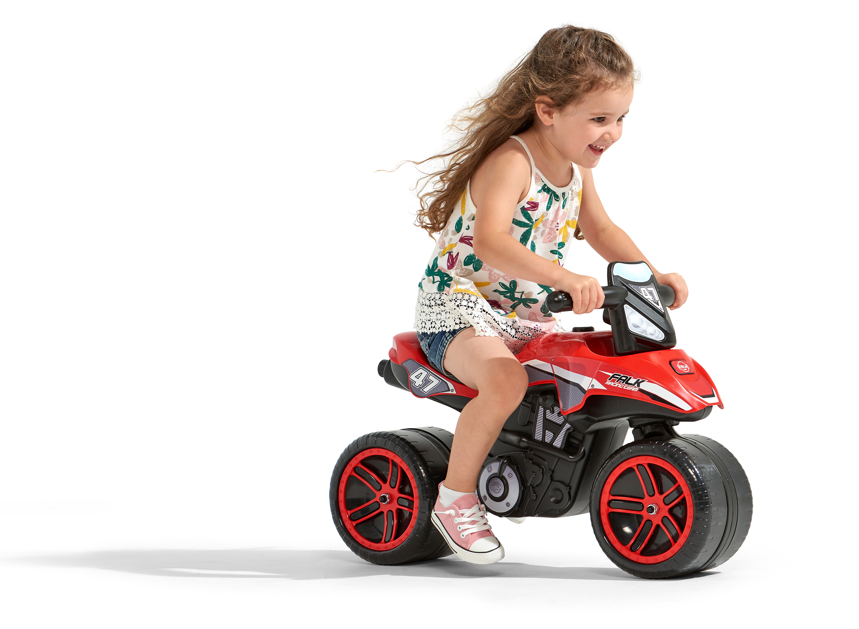 Ein Mädchen fährt lächelnd ein rotes Spielzeugmotorrad. Das Motorrad steht vor weißem Hintergrund. Sie trägt ein Oberteil mit Blumenmuster.