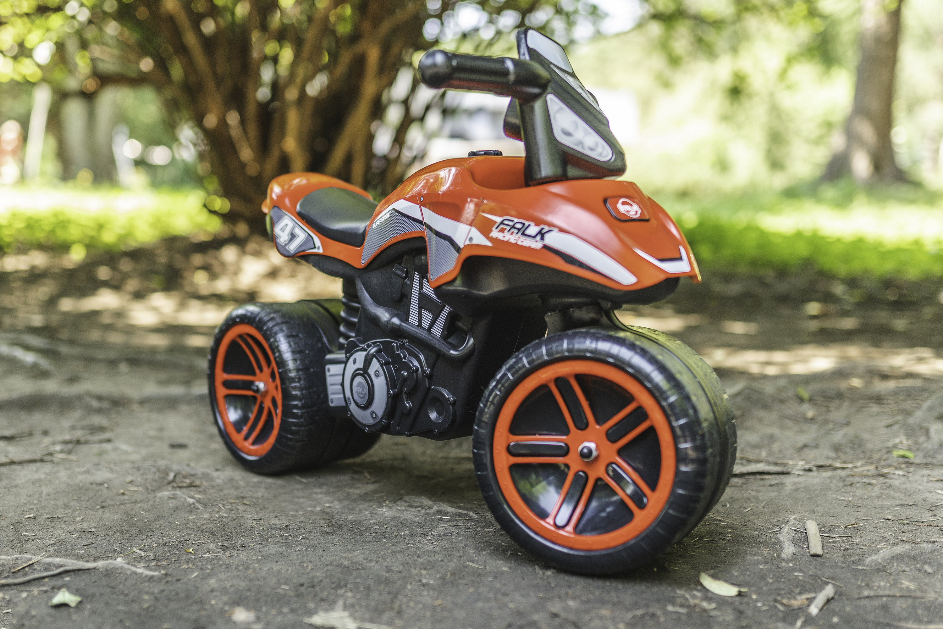 Ein orange-schwarzes Spielzeugmotorrad mit der Nummer 47 steht auf einem Feldweg mit Bäumen im Hintergrund.