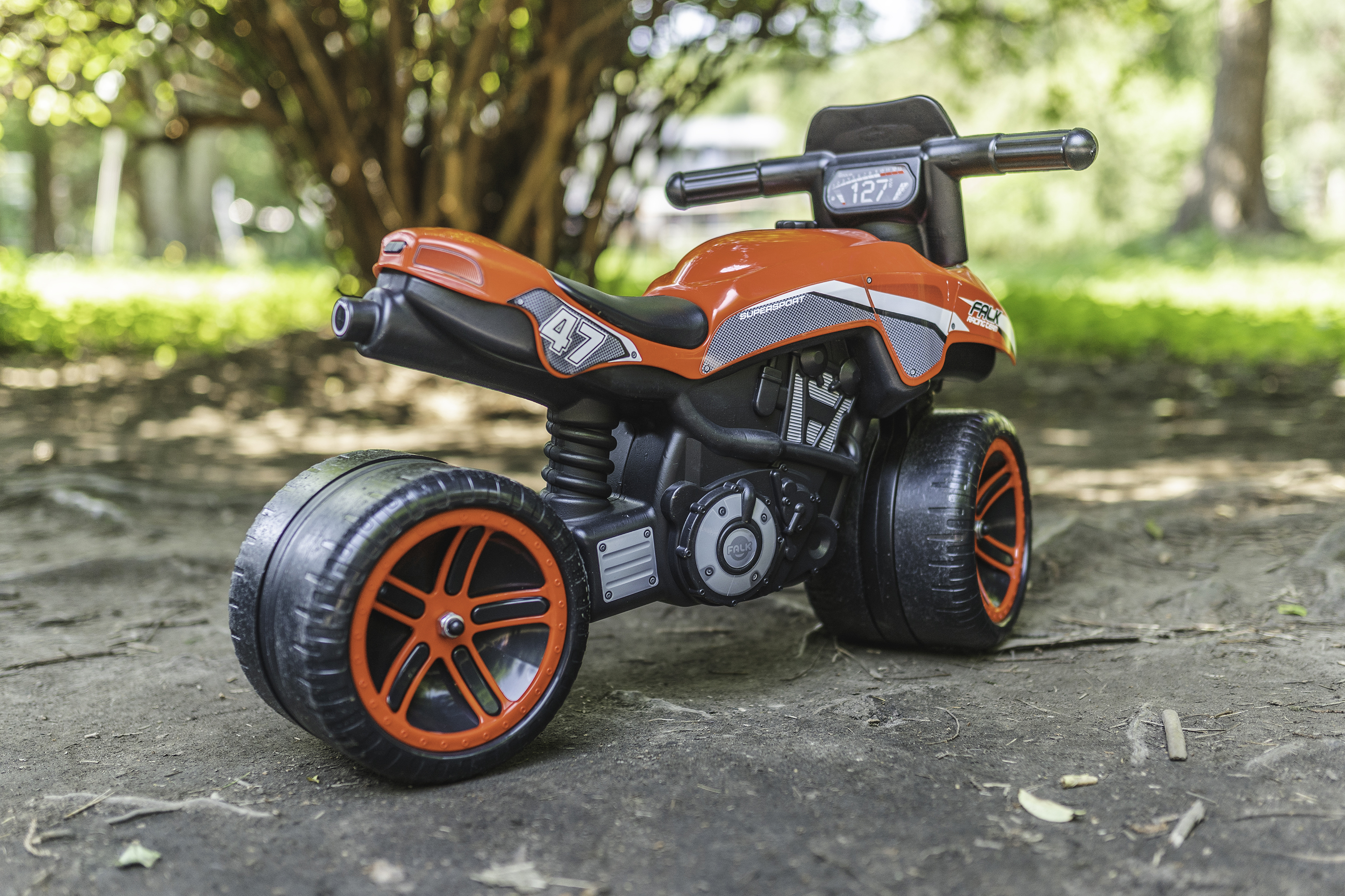Ein orange-schwarzes Spielzeugmotorrad mit der Nummer 47 an der Seite, geparkt auf einem Feldweg mit Bäumen im Hintergrund.
