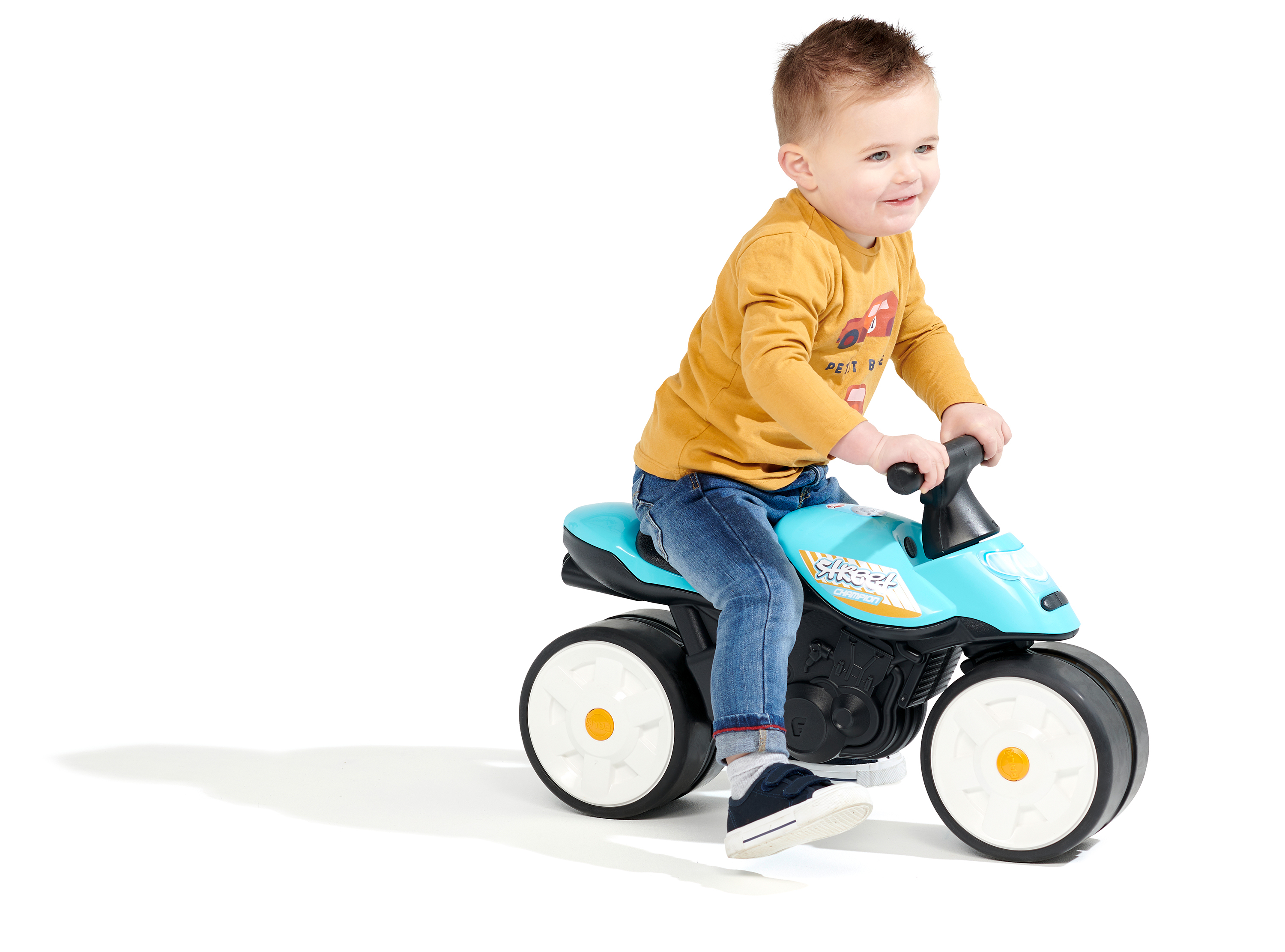 Un bambino piccolo guida una moto giocattolo blu. Indossa jeans e una camicia gialla. Lo sfondo è bianco.