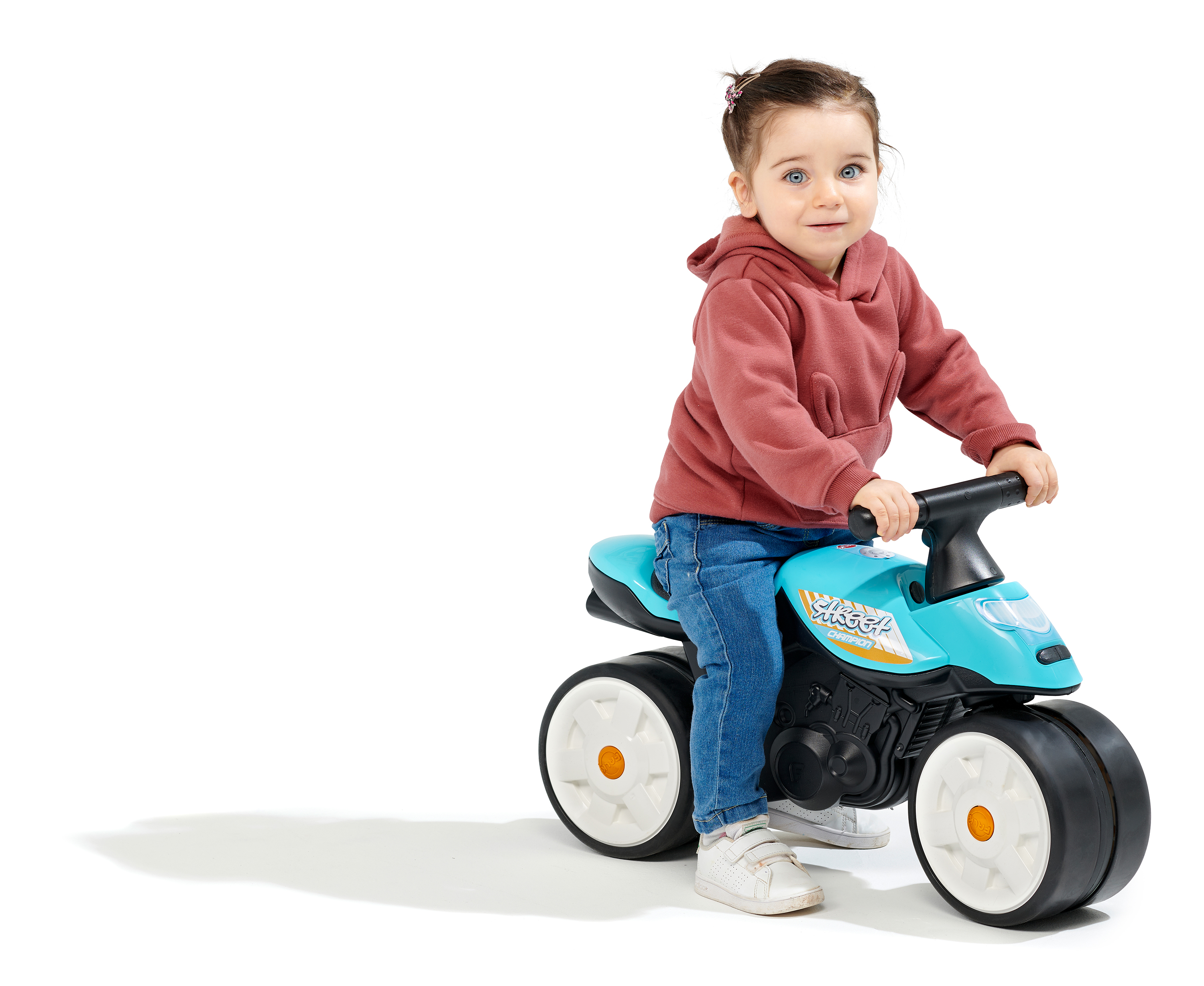 Un bambino piccolo su una moto giocattolo blu, indossa una felpa rossa e jeans, sorride alla fotocamera, sfondo bianco.
