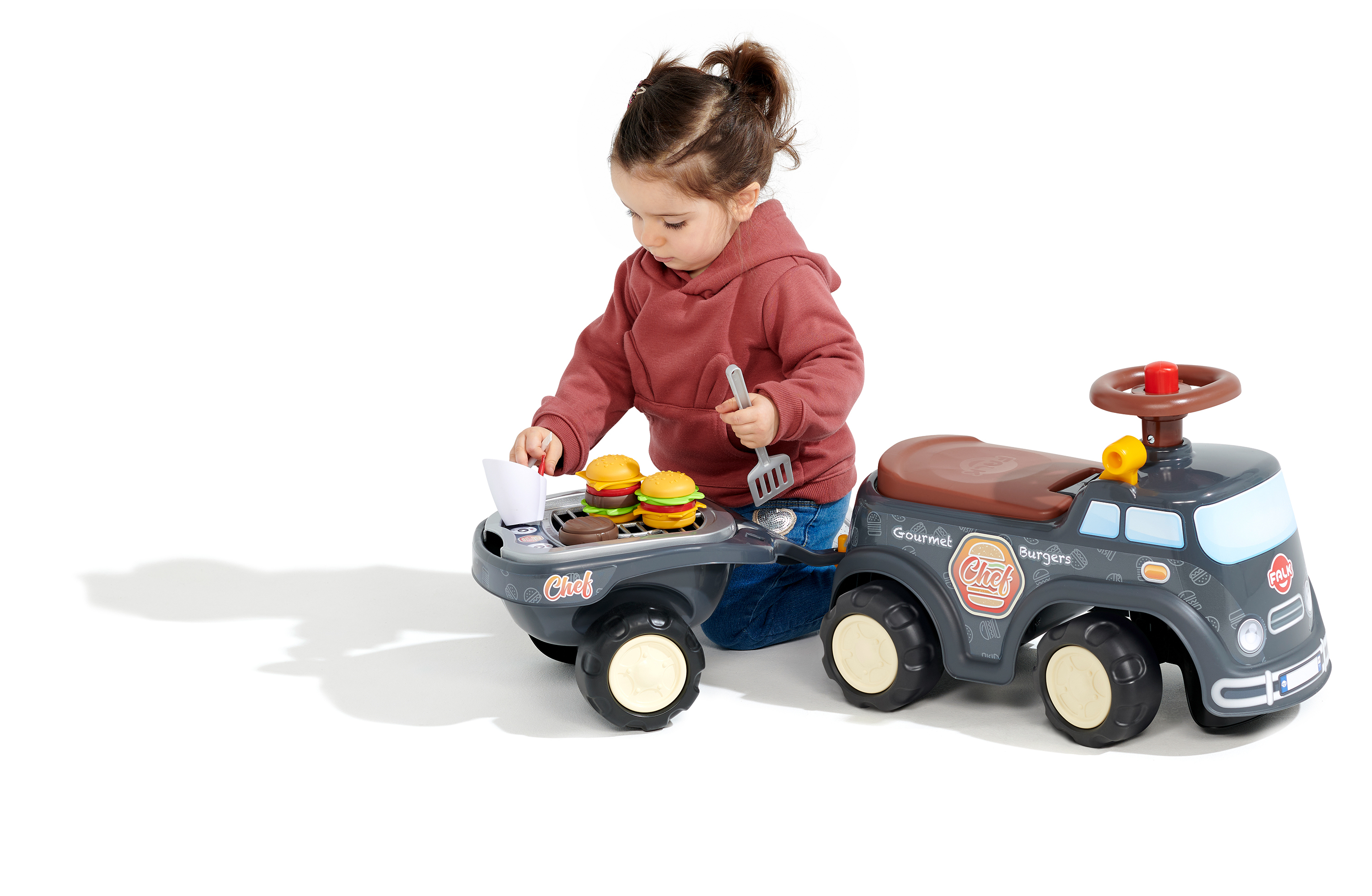 Ein Kind spielt mit einem Spielzeug-Burger-Truck und Grill. Das Kind trägt einen roten Kapuzenpullover und ist auf weißem Grund.