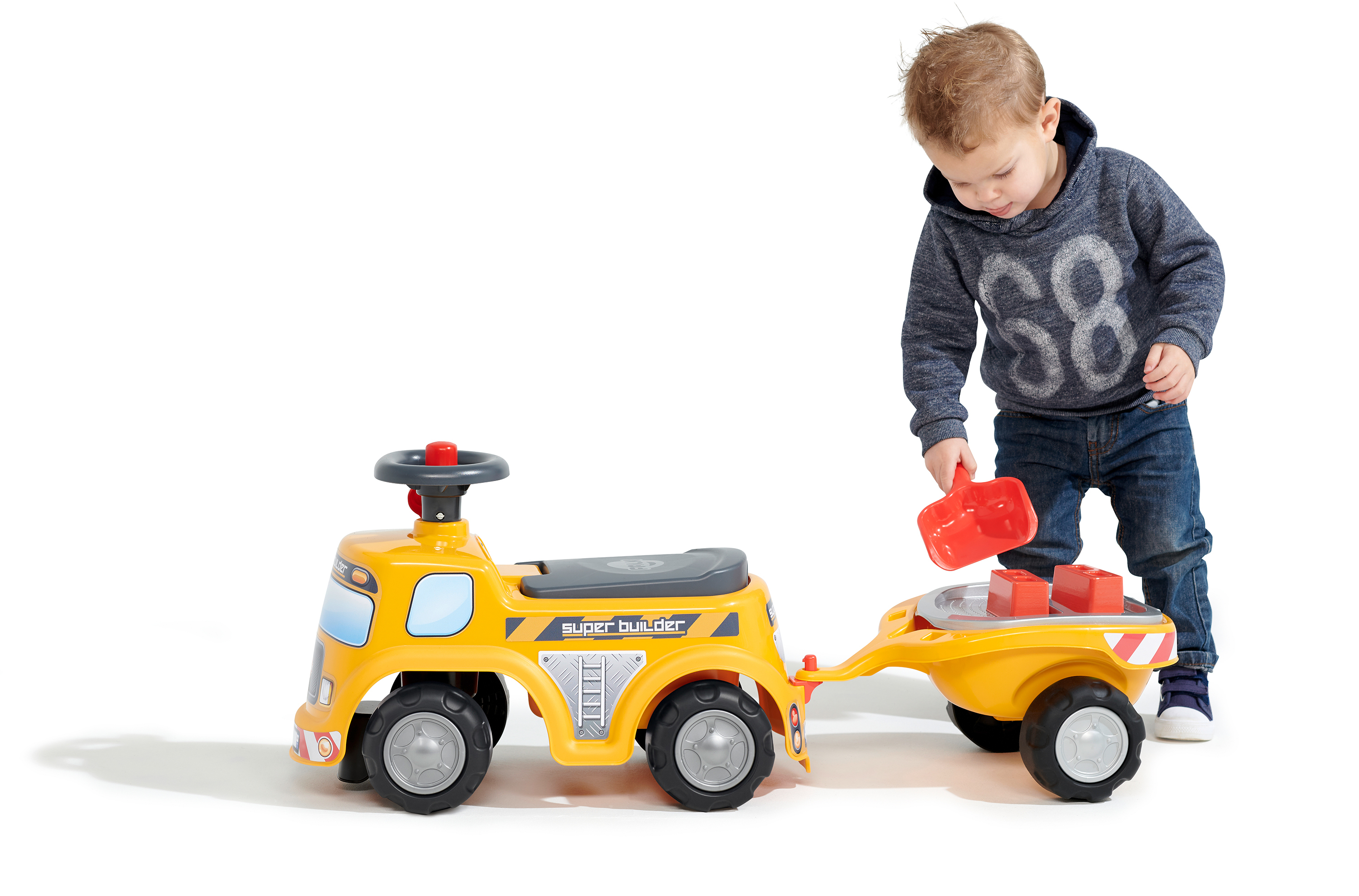 Un bambino gioca con un camion e un rimorchio giocattolo gialli. Il bambino tiene una pala rossa, vicino a blocchi rossi.