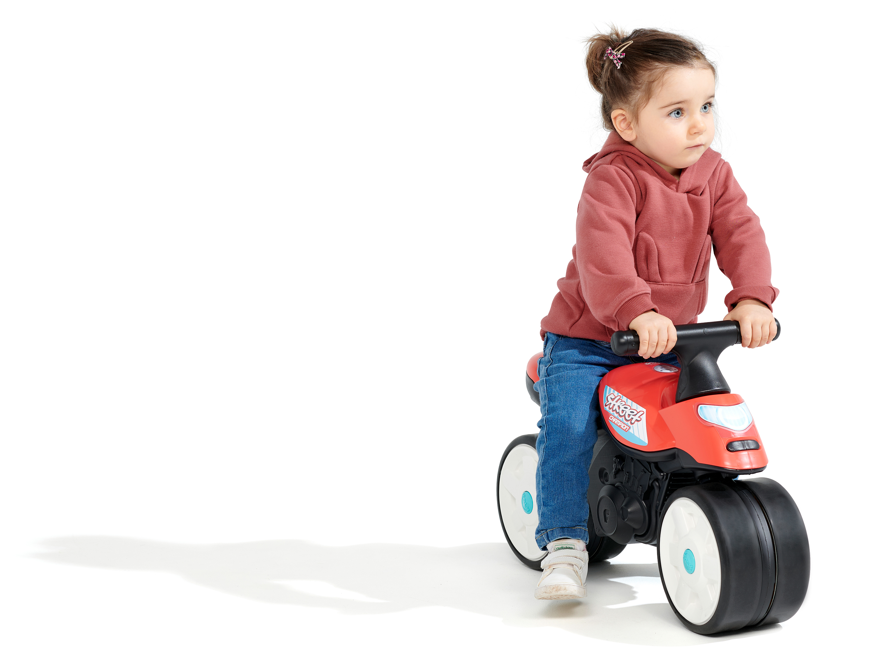 Ein junges Kind auf einem roten Spielzeugmotorrad, trägt einen roten Kapuzenpullover, Jeans und weiße Schuhe, vor weißem Hintergrund.