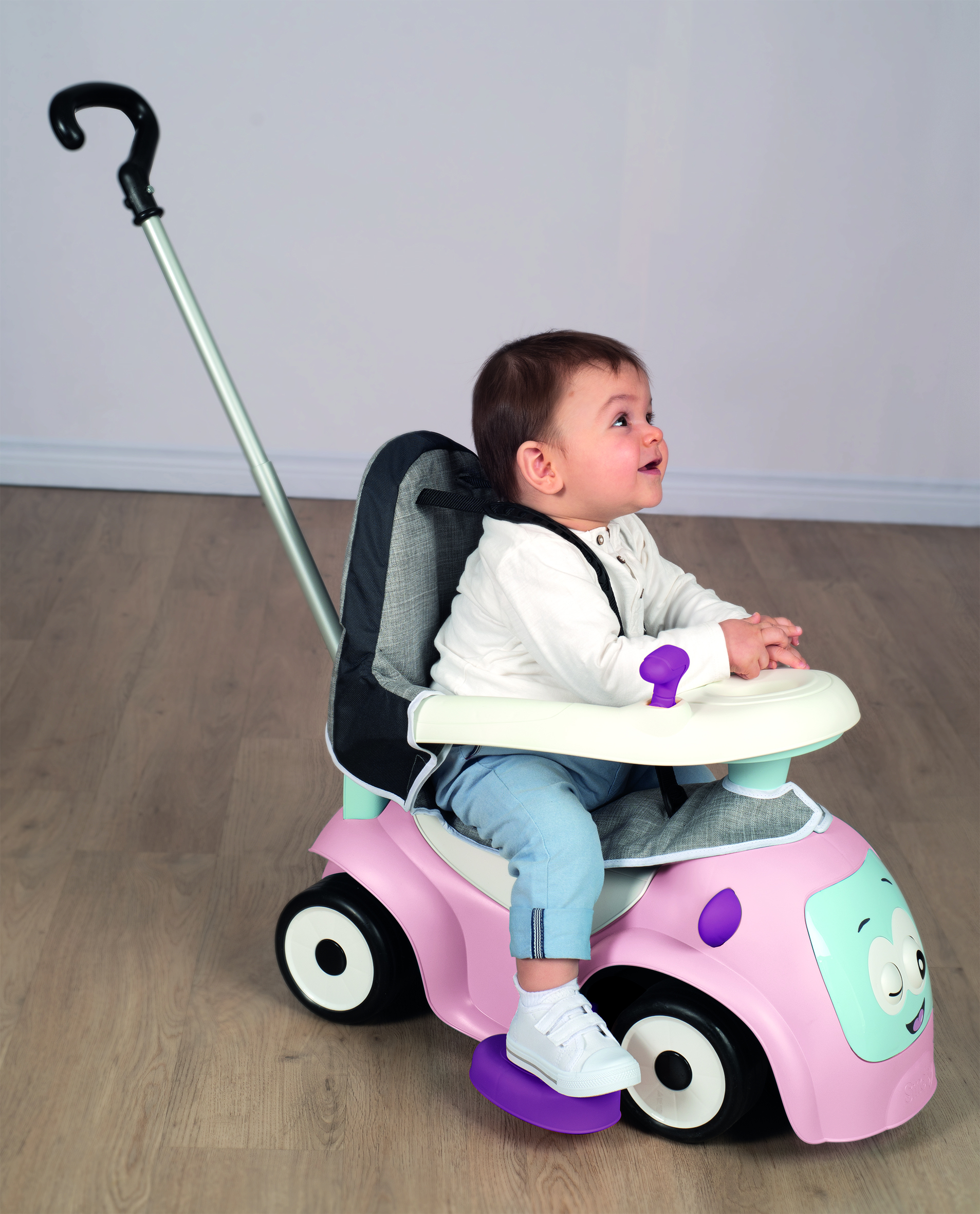 Ein Baby sitzt in einem rosa Spielzeugauto und blickt nach oben. Das Auto hat einen Schiebegriff. Der Hintergrund ist eine helle Wand und ein Holzboden.