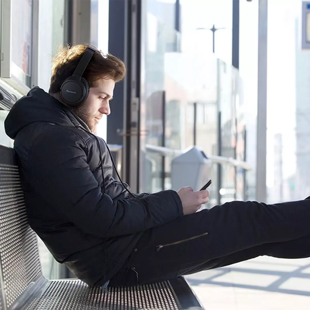 Ein Mann mit Kopfhörern sitzt auf einer Bank und schaut auf sein Handy. Er trägt eine schwarze Jacke und Hose. Im Hintergrund ist ein modernes Gebäude zu sehen.