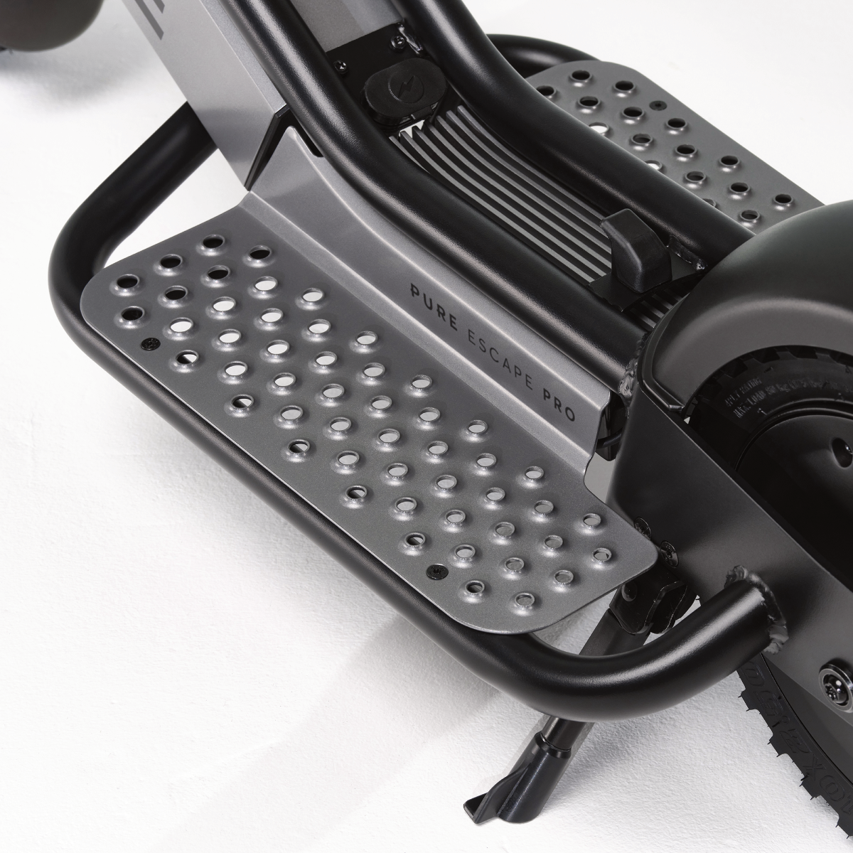 Nahaufnahme der Fußstütze eines Rollers mit dem Text 'PURE ESCAPE PRO'. Die Fußstütze ist grau mit kreisförmigen Löchern und einem schwarzen Rahmen.