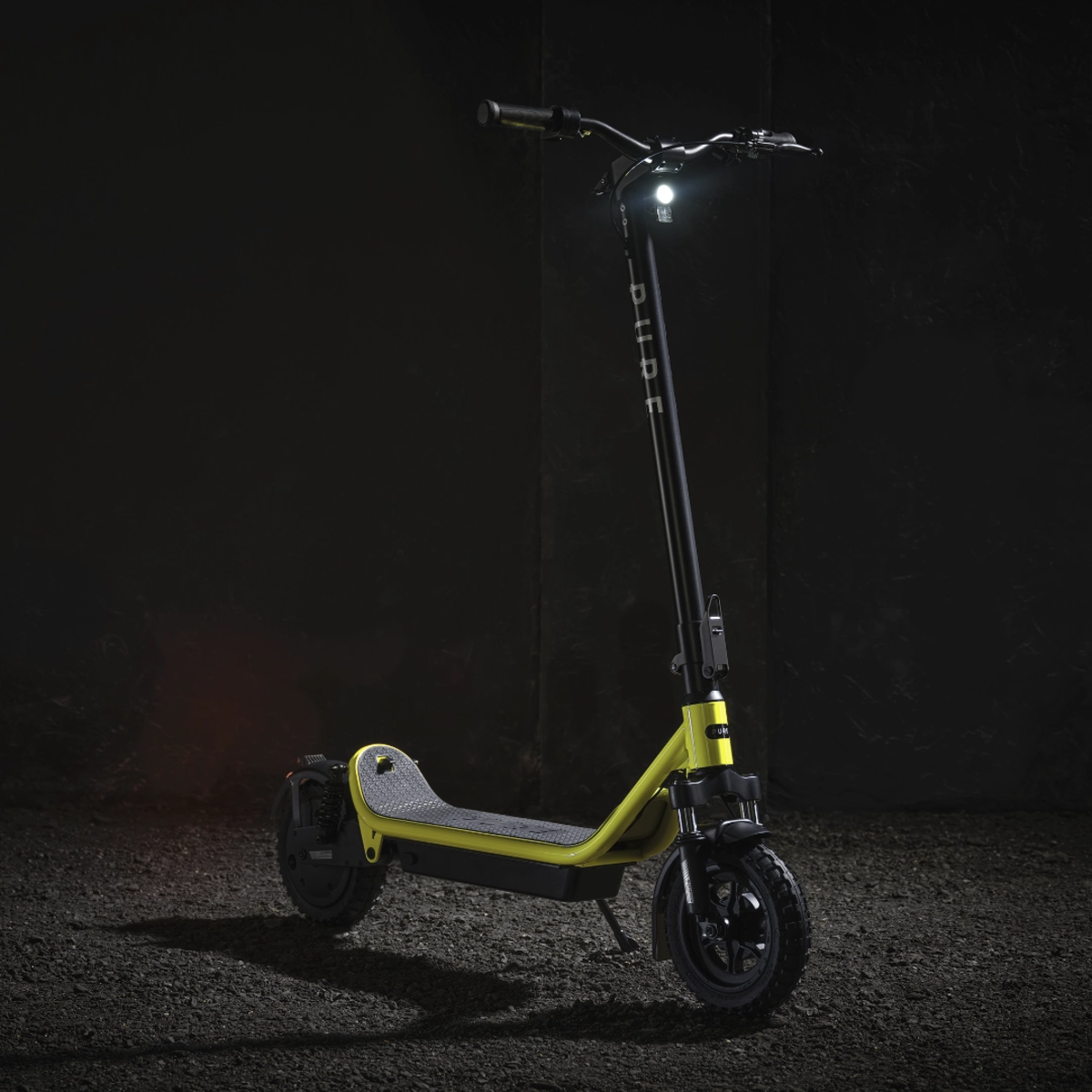 Ein gelb-schwarzer Elektroroller steht auf einer strukturierten Oberfläche. Der Scheinwerfer des Rollers ist eingeschaltet und leuchtet die Front aus.