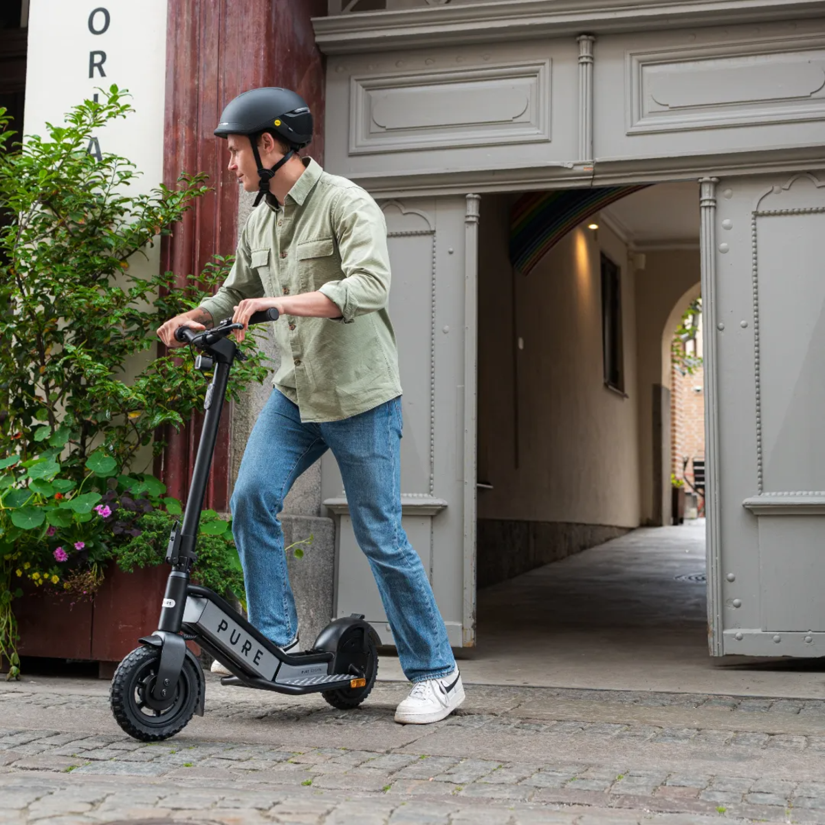 Ein Mann mit Helm fährt mit einem schwarzen Roller aus einer Tür. Er trägt Jeans und ein grünes Hemd. Auf dem Roller steht 'PURE'.
