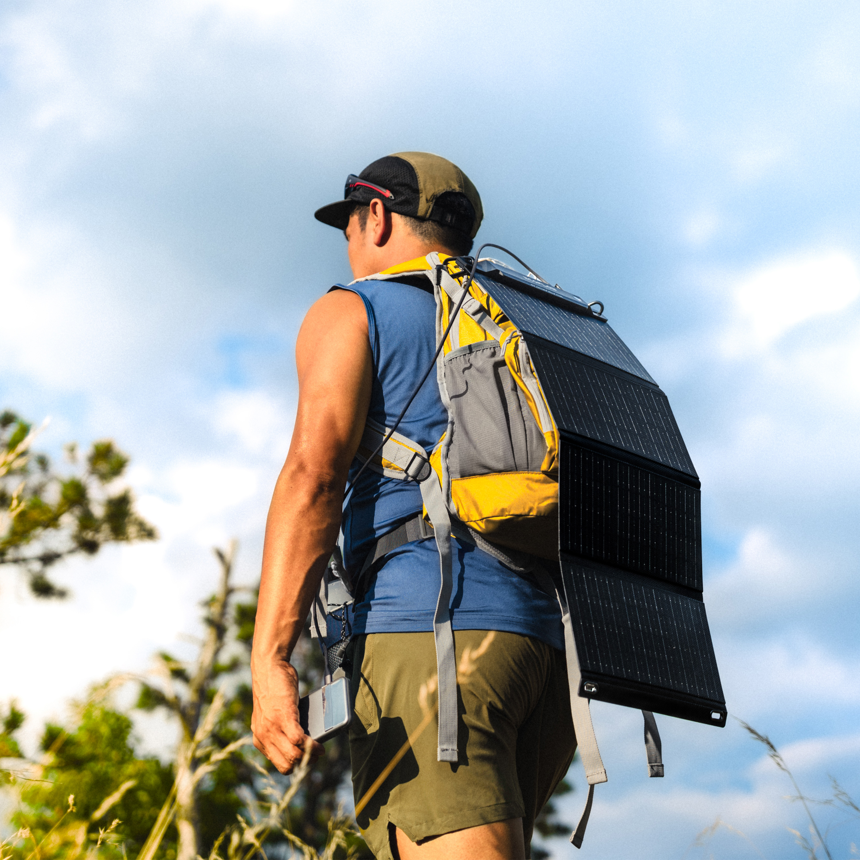 Un hombre con una mochila con panel solar camina al aire libre. El cielo es azul con nubes. Lleva gorra y pantalones cortos.