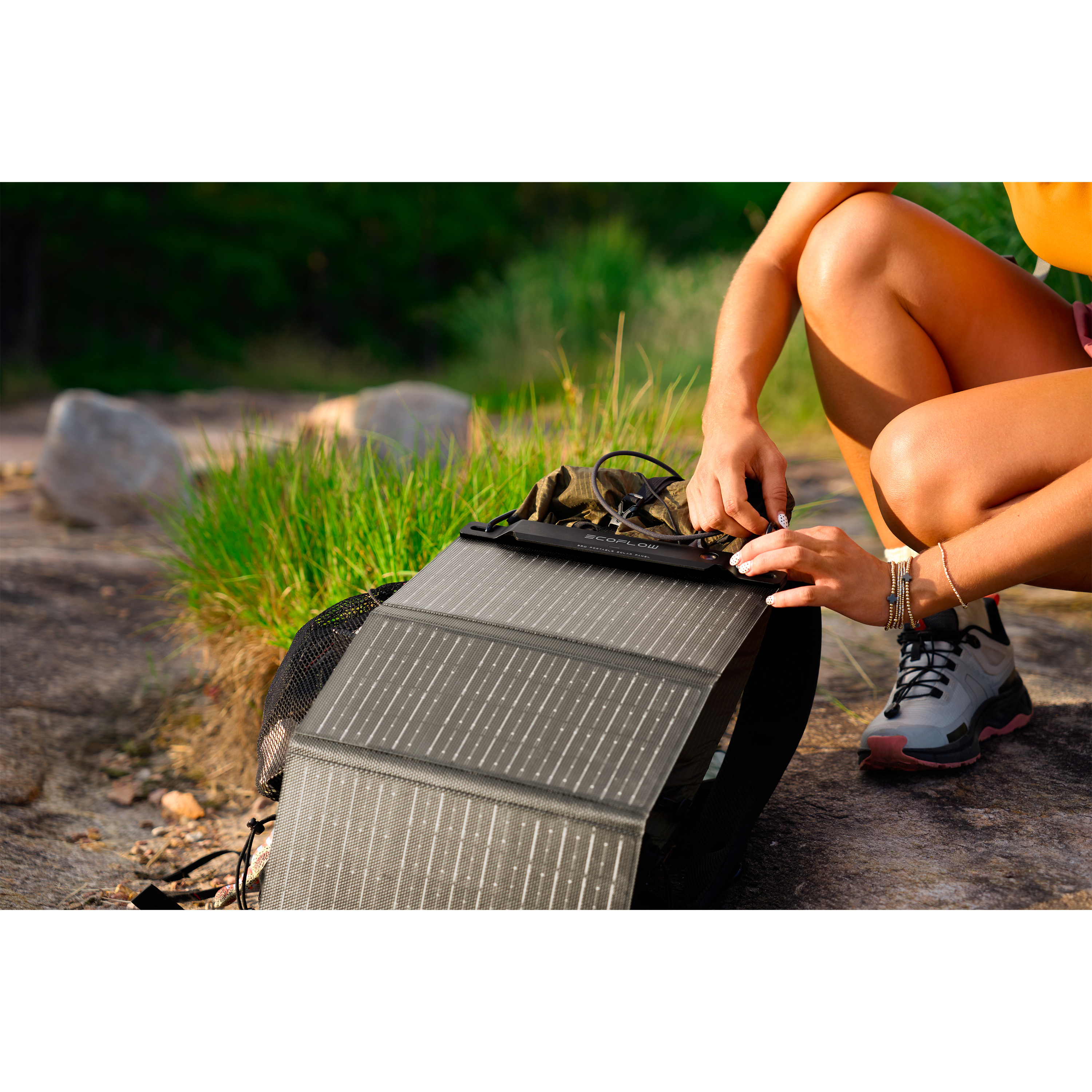Una persona está arrodillada, ajustando un panel solar sobre una roca. Hierba verde y una mochila en el fondo.