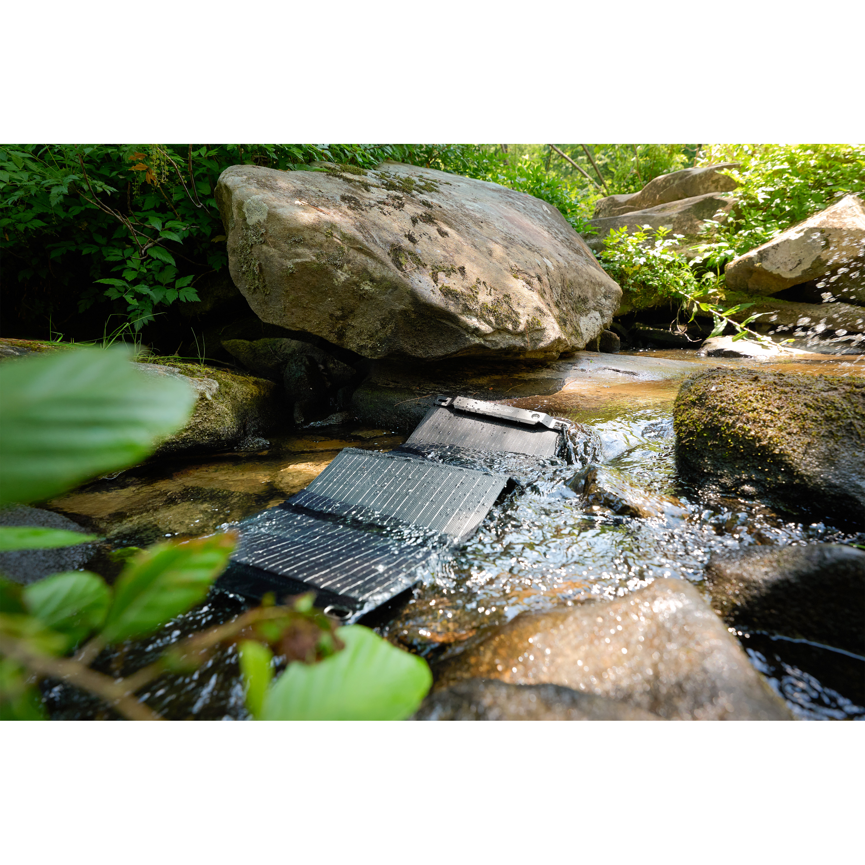 Un panel solar en un arroyo. Rocas lo rodean. El agua fluye sobre él. Follaje verde en el fondo.