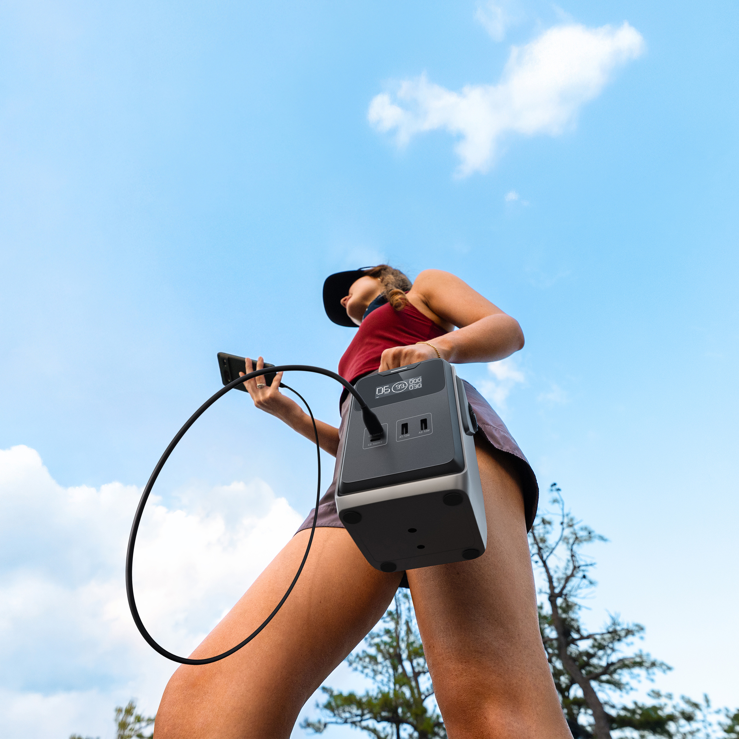Eine Person hält eine Powerstation und ein Telefon vor blauem Himmel. Die Powerstation hat einen Bildschirm und Anschlüsse.