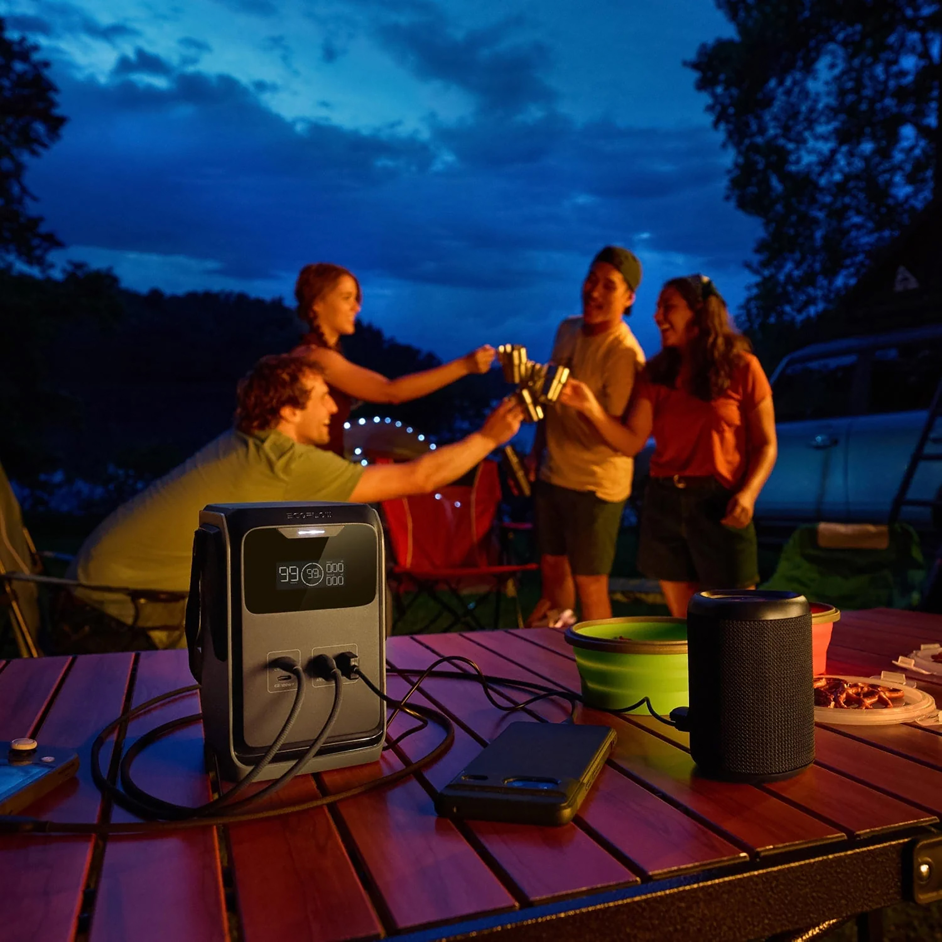 Leute stoßen sich am Campingplatz bei Dämmerung mit Getränken zu. Eine Powerstation, ein Lautsprecher und ein Telefon liegen auf dem Tisch.