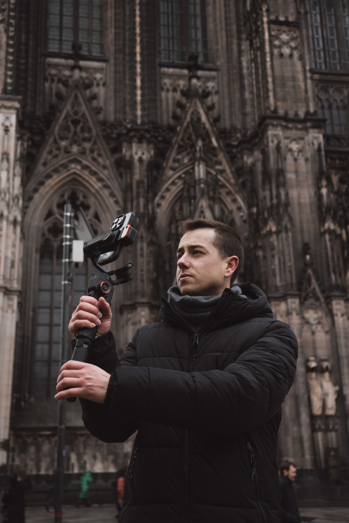 Mężczyzna w czarnym płaszczu trzyma stabilizator kamery przed katedrą.