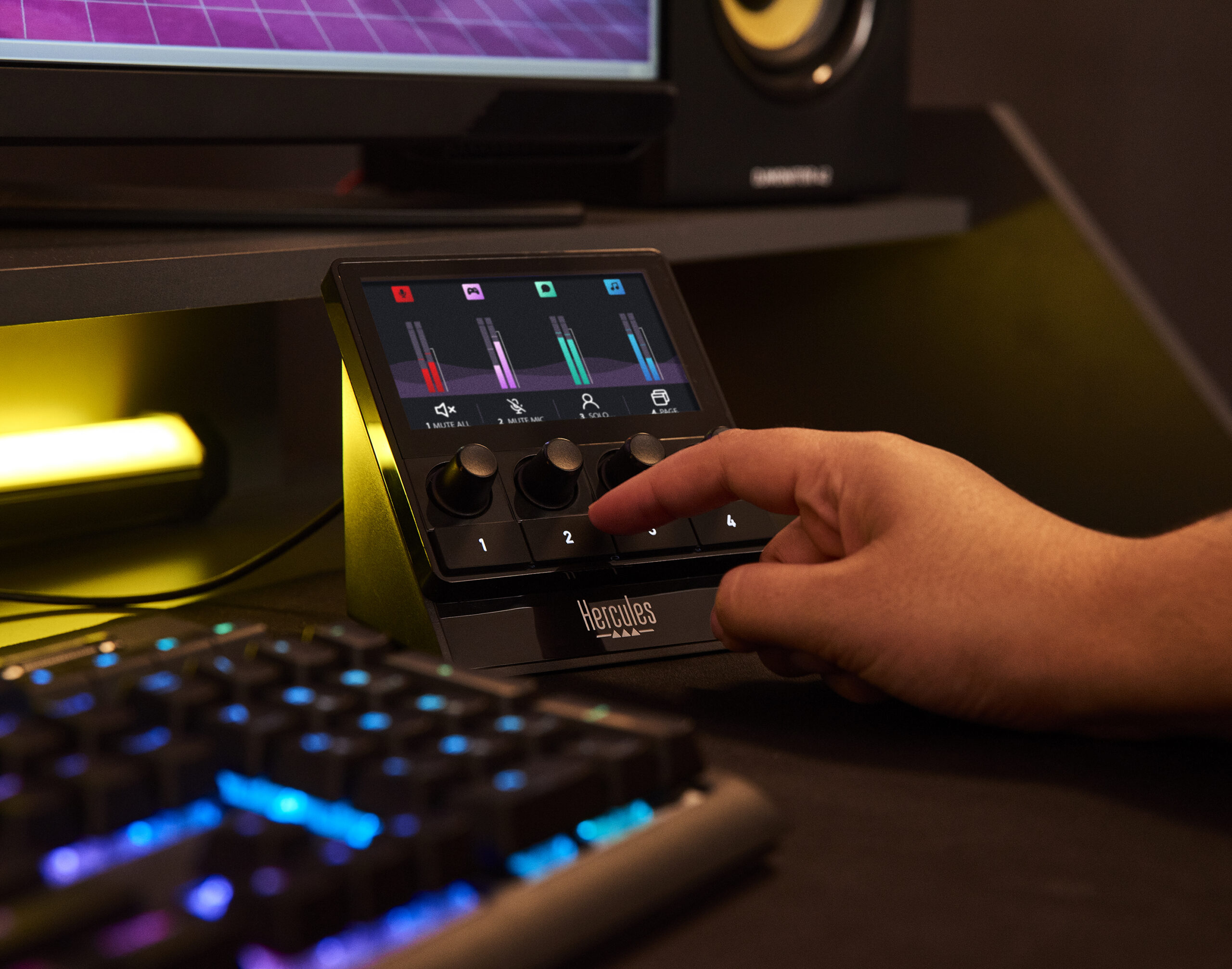 Een hand raakt een Hercules audiocontroller aan. Display toont audioniveaus. Toetsenbord op de voorgrond, monitor en luidspreker op de achtergrond.