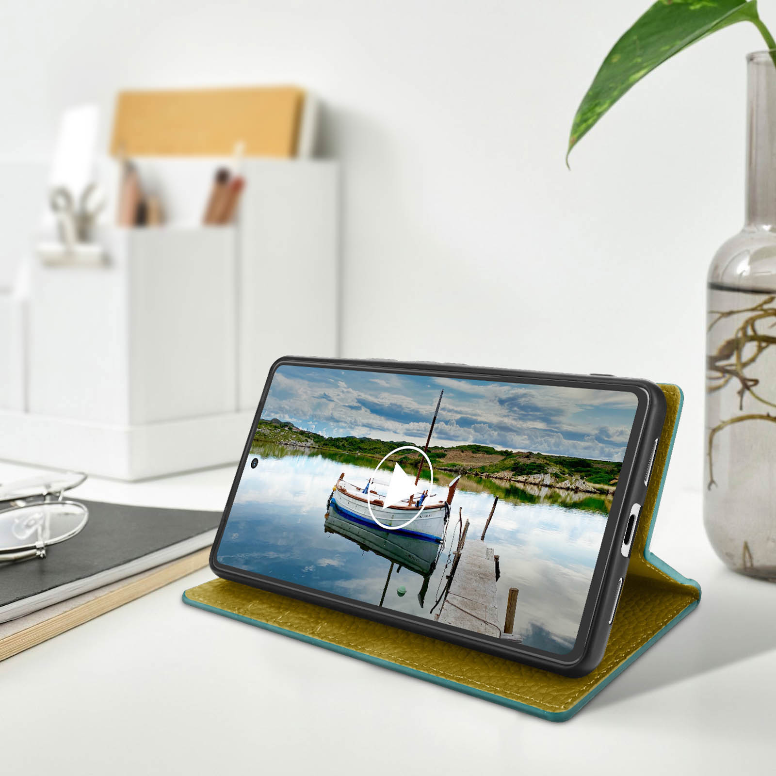 Een telefoon toont een boot op het water. De telefoon staat op een bureau met een plant, bril en notitieboek.