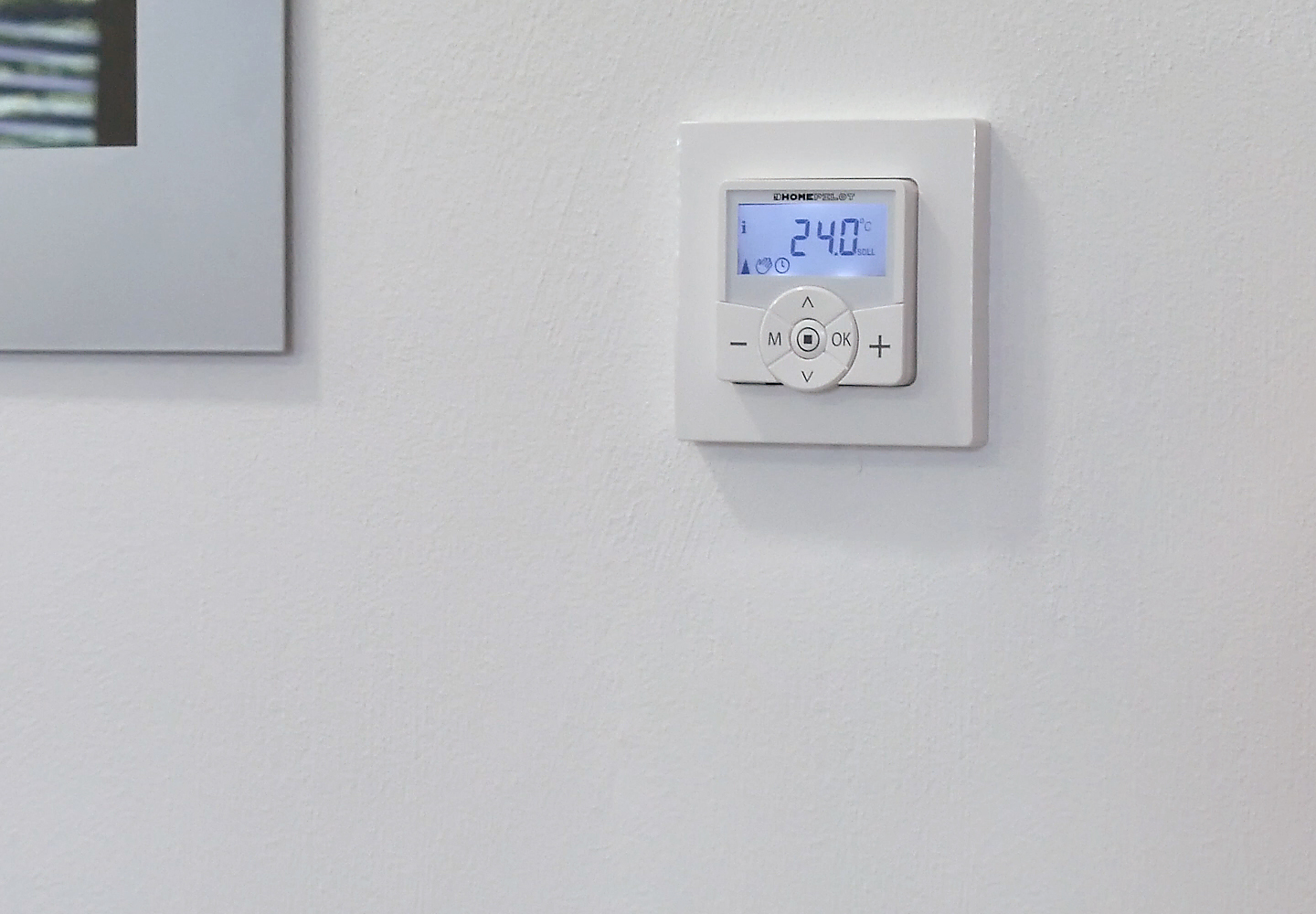 Eine weiße Wand mit einem Thermostat, der 24,0 Grad Celsius anzeigt. Auf dem Thermostat sind Knöpfe sichtbar. Links ist ein silberner Rahmen.