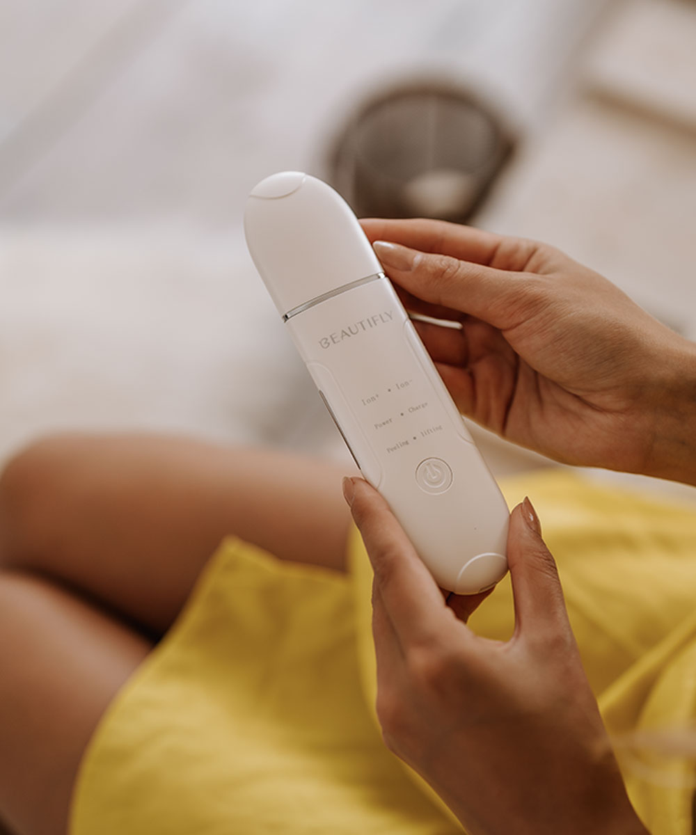 Osoba trzyma białe urządzenie do pielęgnacji skóry 'BEAUTIFLY'. Osoba ma na sobie żółtą górę. Tło jest rozmyte.