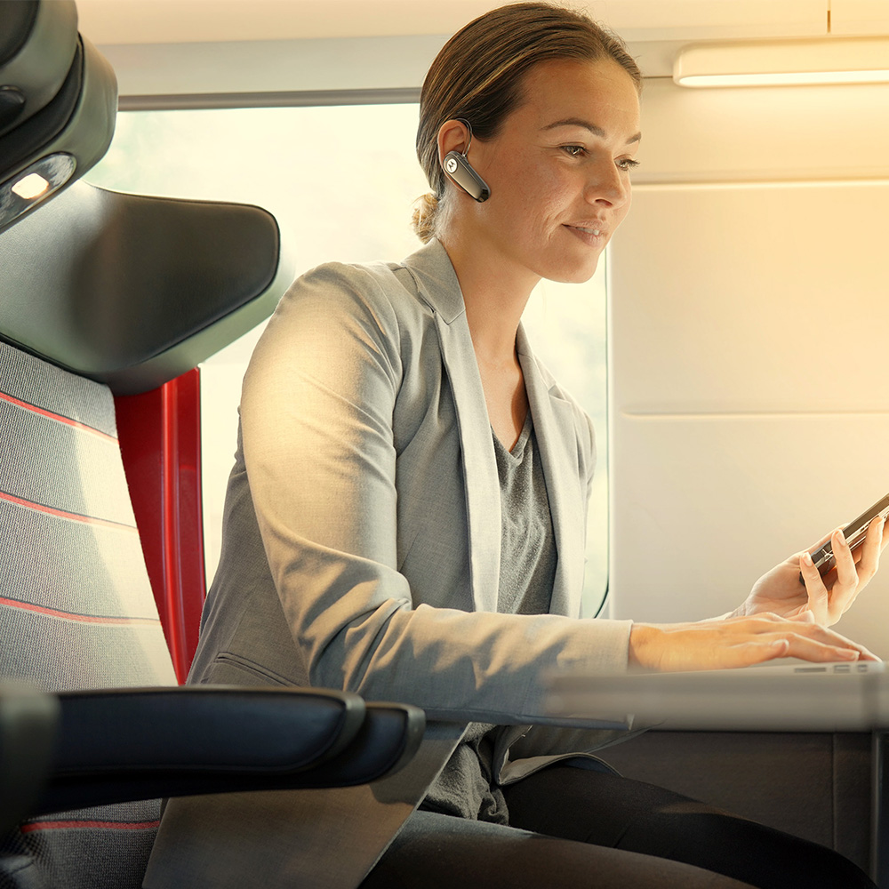 Frau im Zug mit Laptop und Telefon, trägt ein Headset. Sie trägt einen grauen Blazer. Zuginnenraum sichtbar.
