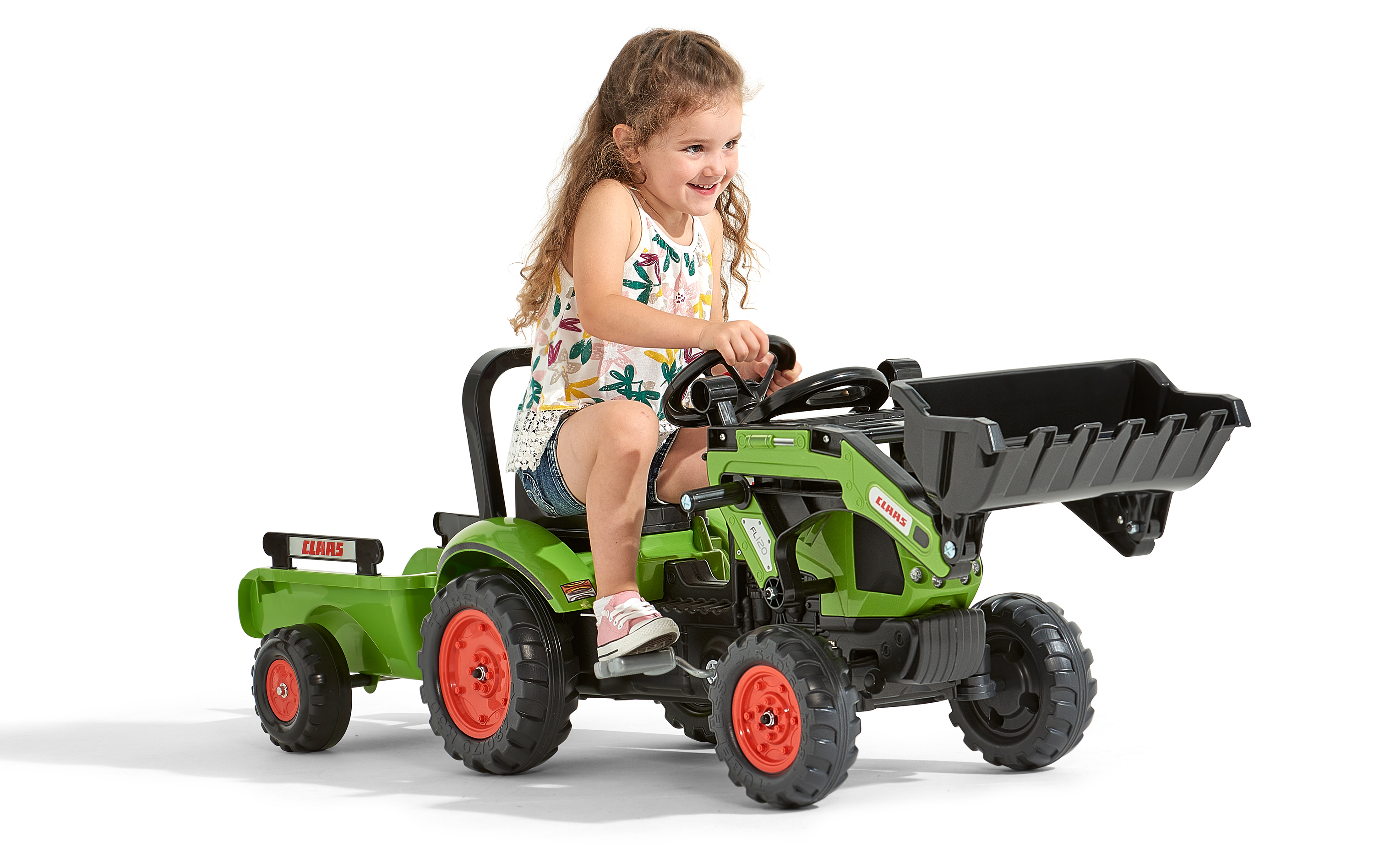 Ein lächelndes Kind auf einem grünen Spielzeugtraktor. Der Traktor hat einen Frontlader und einen Anhänger. Weißer Hintergrund.