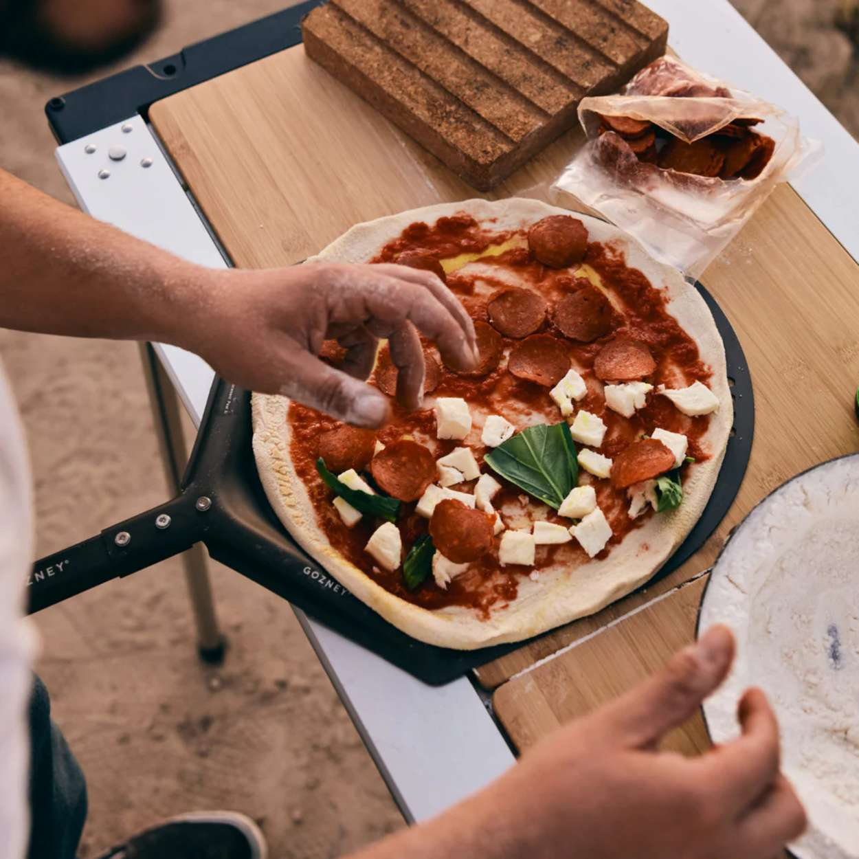 Eine Person legt Peperoni auf eine Pizza. Die Pizza hat Soße, Käse und Basilikum. Zutaten liegen auf einer Holzoberfläche.