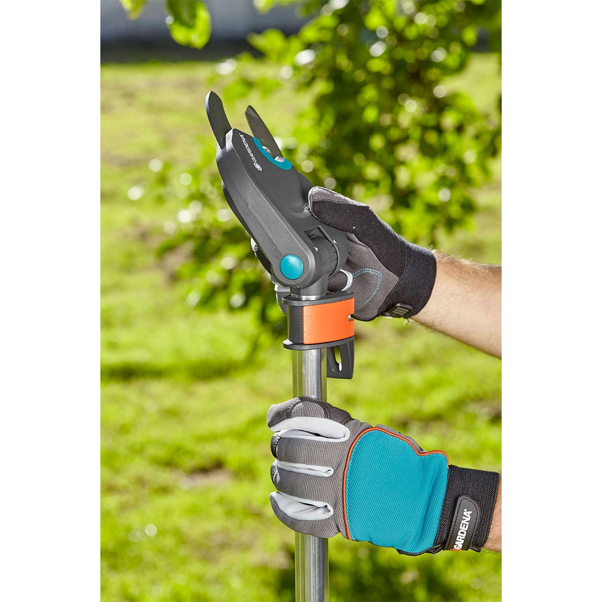 Hände mit Handschuhen halten ein Gartengerät. Das Werkzeug ist grau und schwarz mit einem orangenen Teil. Der Hintergrund ist grünes Gras und Bäume.